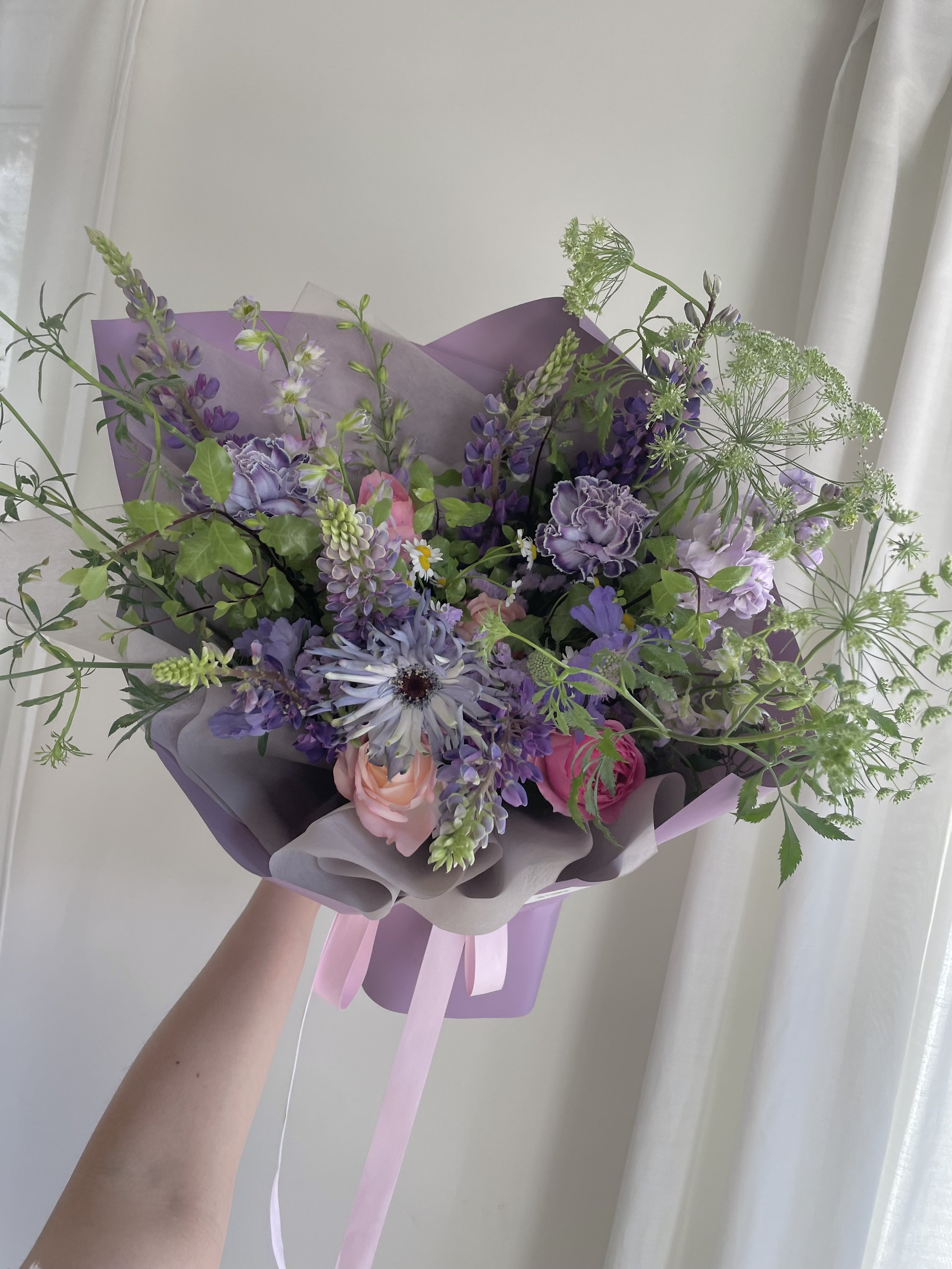 Hand holding a bouquet of purple, pink, and white flowers wrapped in purple and gray paper with pink ribbon, in front of a light-colored wall and white curtains.