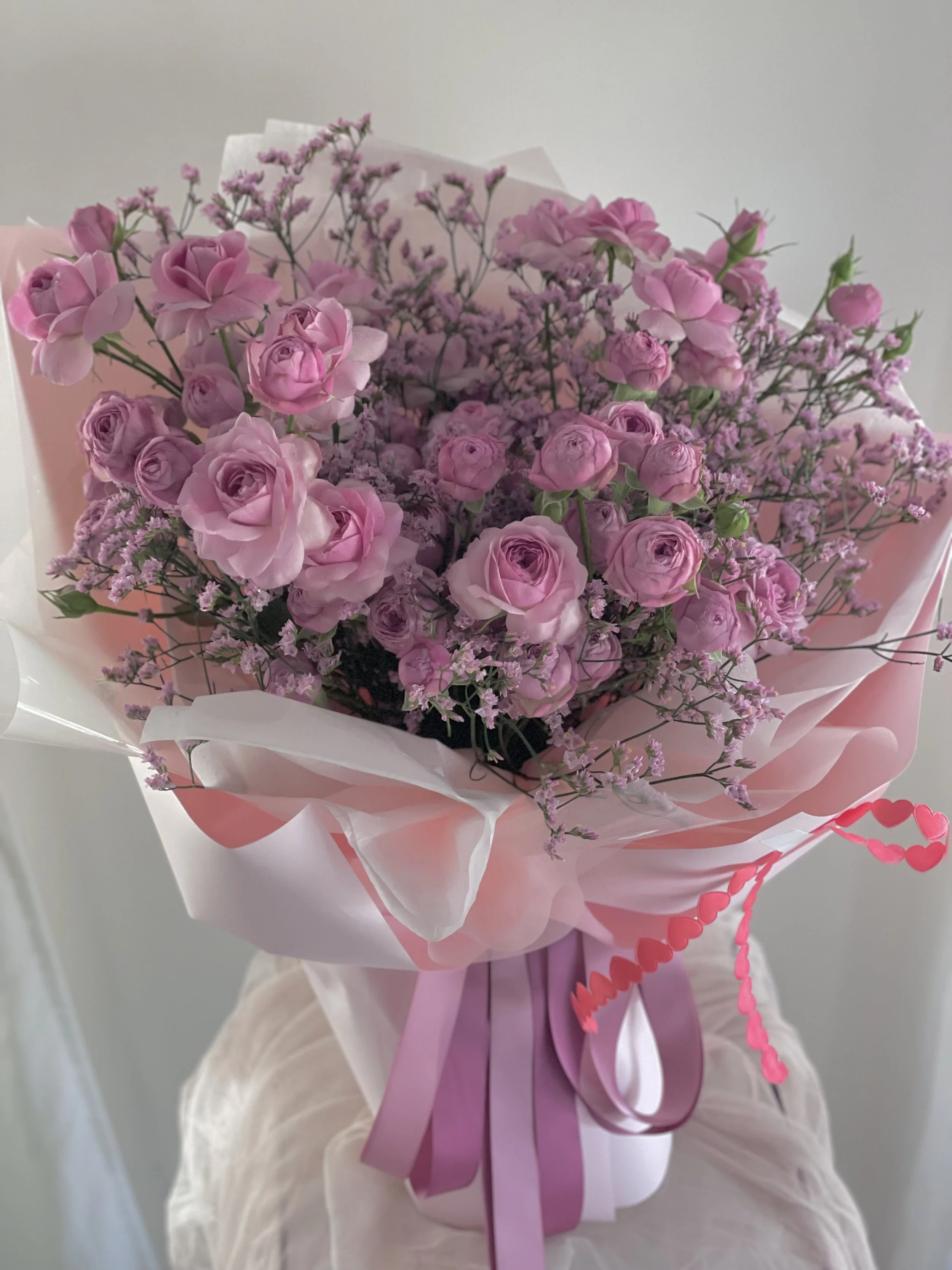 Pink and purple bouquet of roses and small filler flowers wrapped in pink and white paper with pink ribbon, sitting on a white surface.