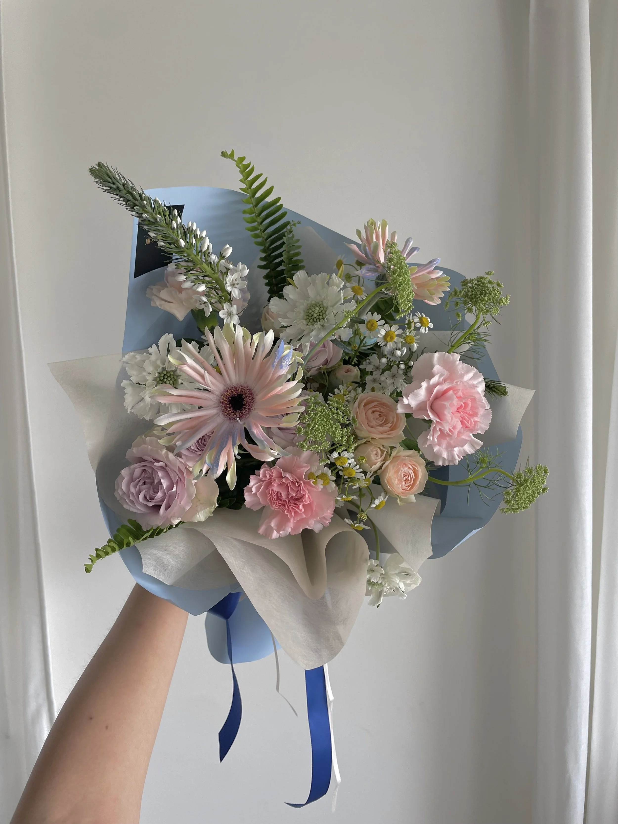 A hand holding a bouquet of pink, white, and cream flowers wrapped in beige and blue paper with blue and white ribbons, against a neutral background.