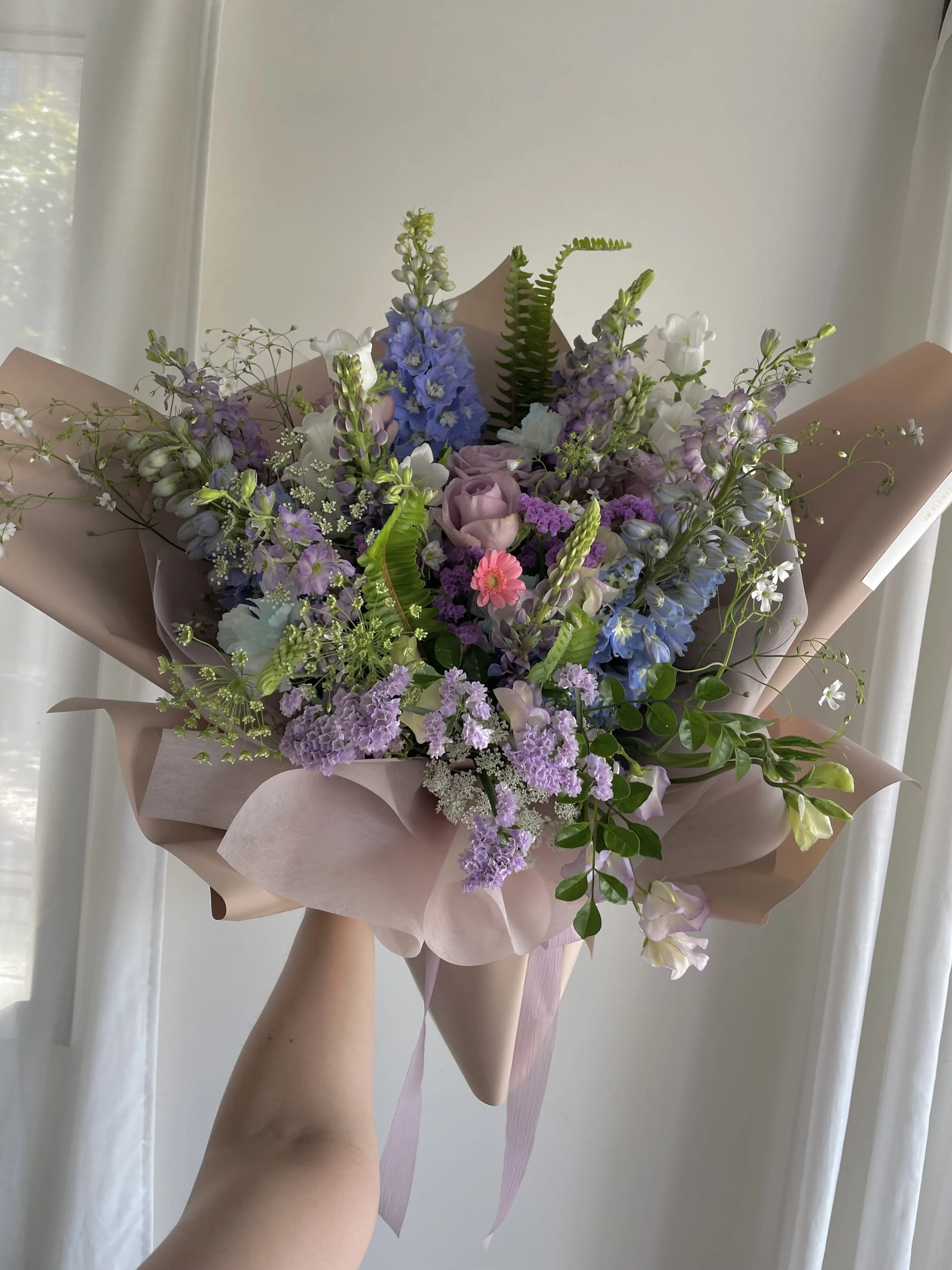 A person holds a bouquet of various purple, pink, and white flowers wrapped in pink paper.