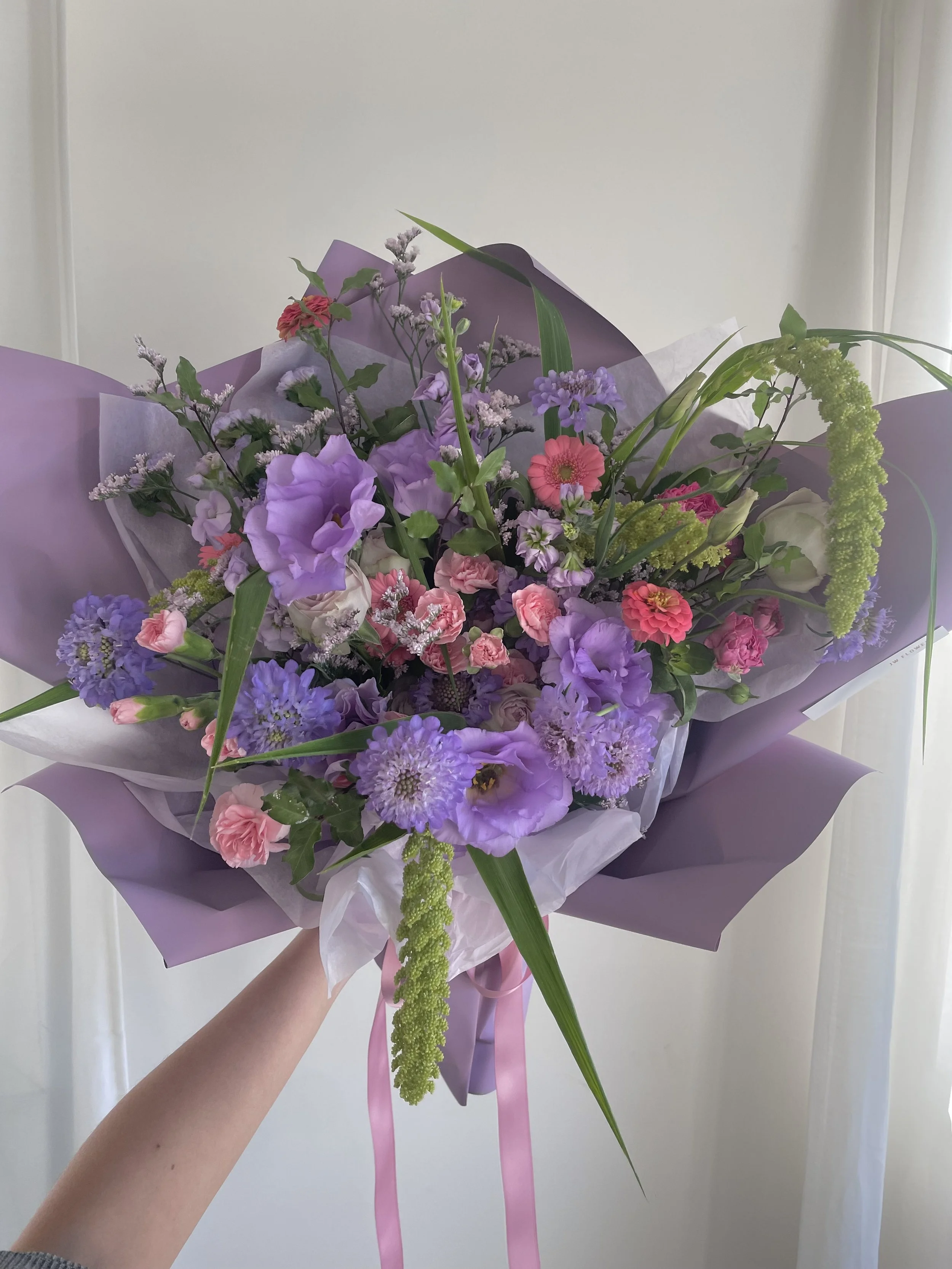 A bouquet of colorful flowers wrapped in purple and white paper, tied with a pink ribbon, held by a person's hand.