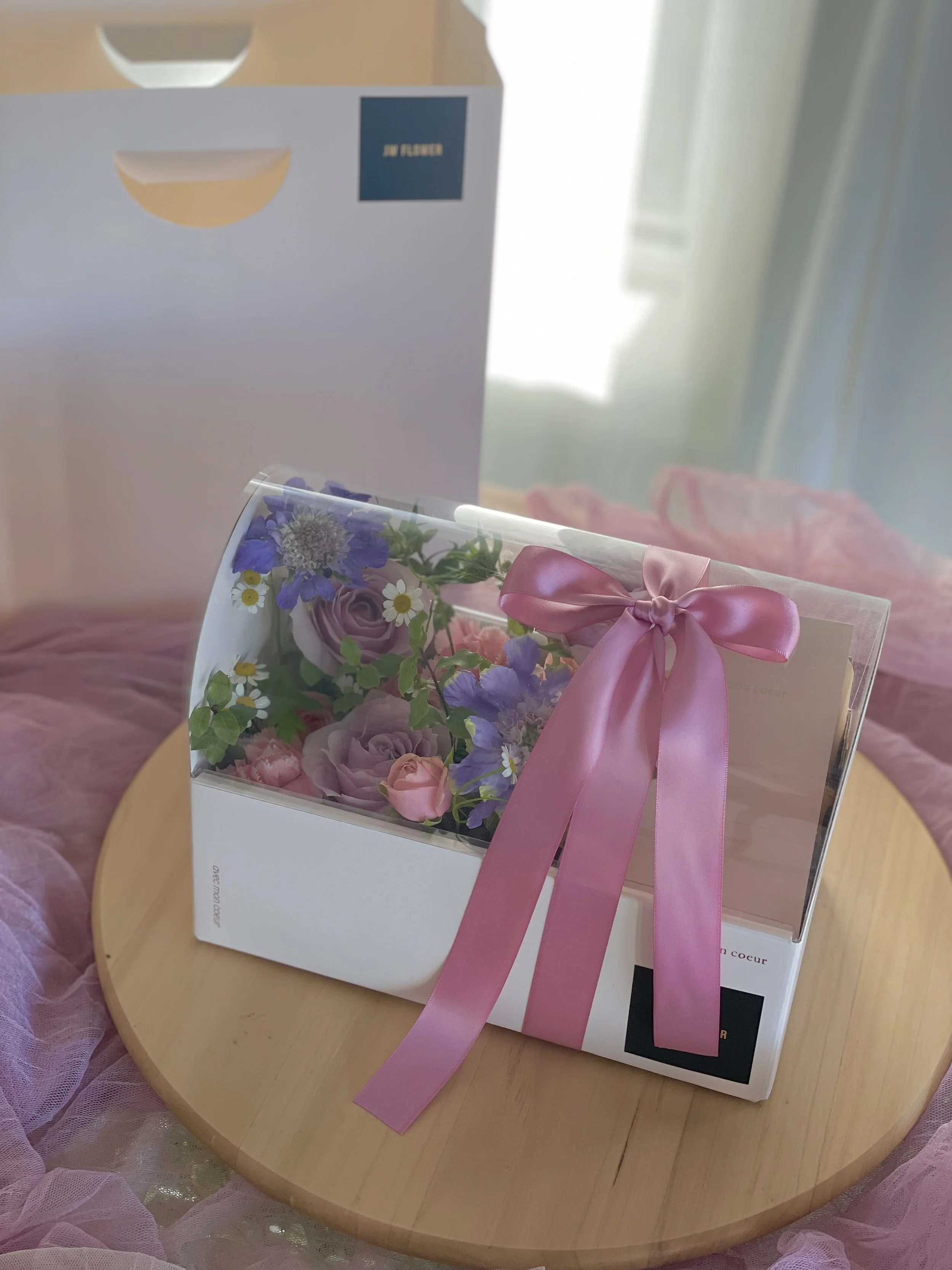 A floral arrangement with purple, pink, and white flowers inside a transparent box, tied with a pink ribbon, sitting on a wooden round table with pink tulle fabric around it.