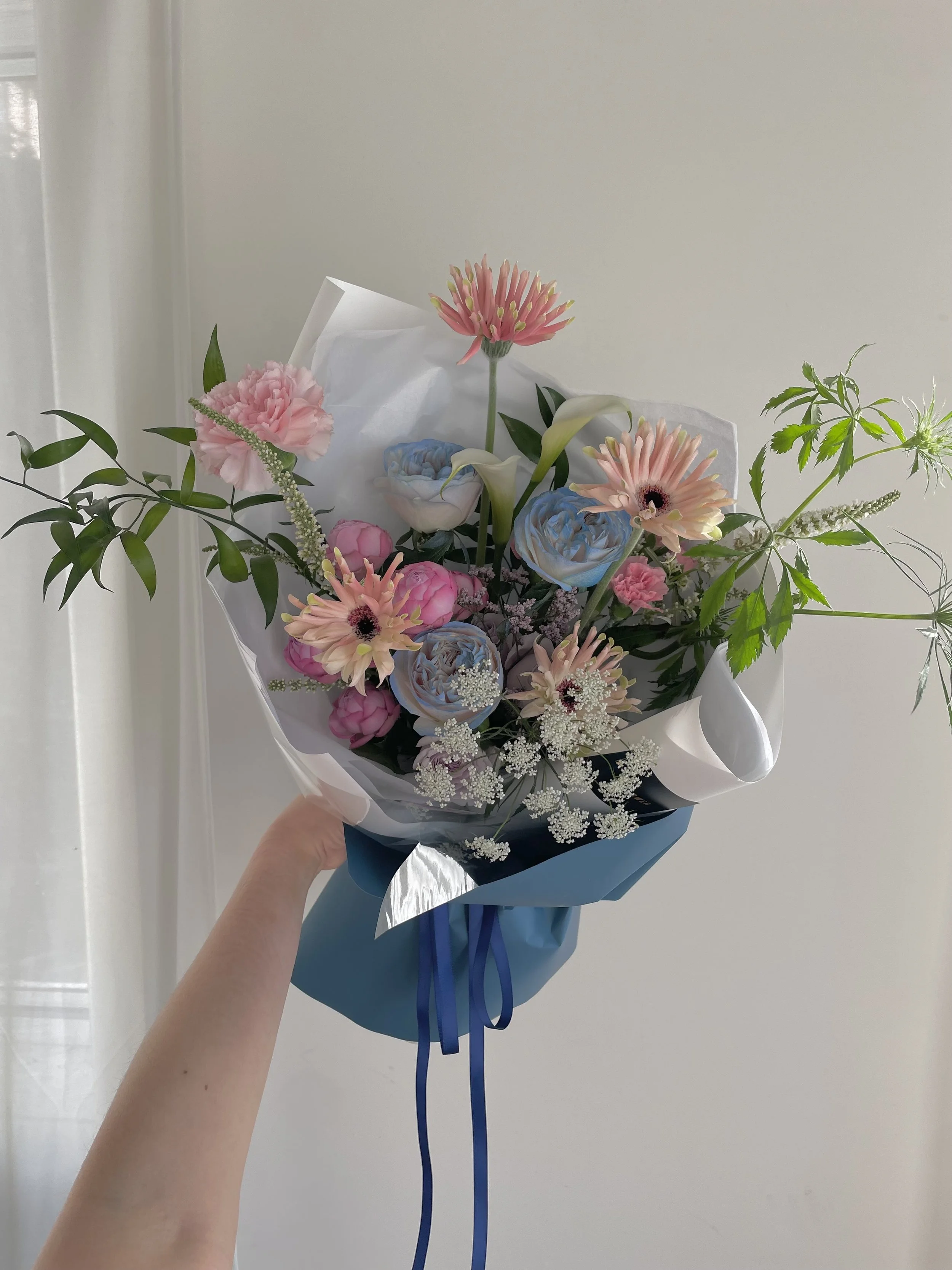 A hand holding a pastel-colored flower bouquet wrapped in blue paper, featuring pink carnations, white calla lilies, blue roses, pink gerbera daisies, and white baby's breath, against a neutral background.