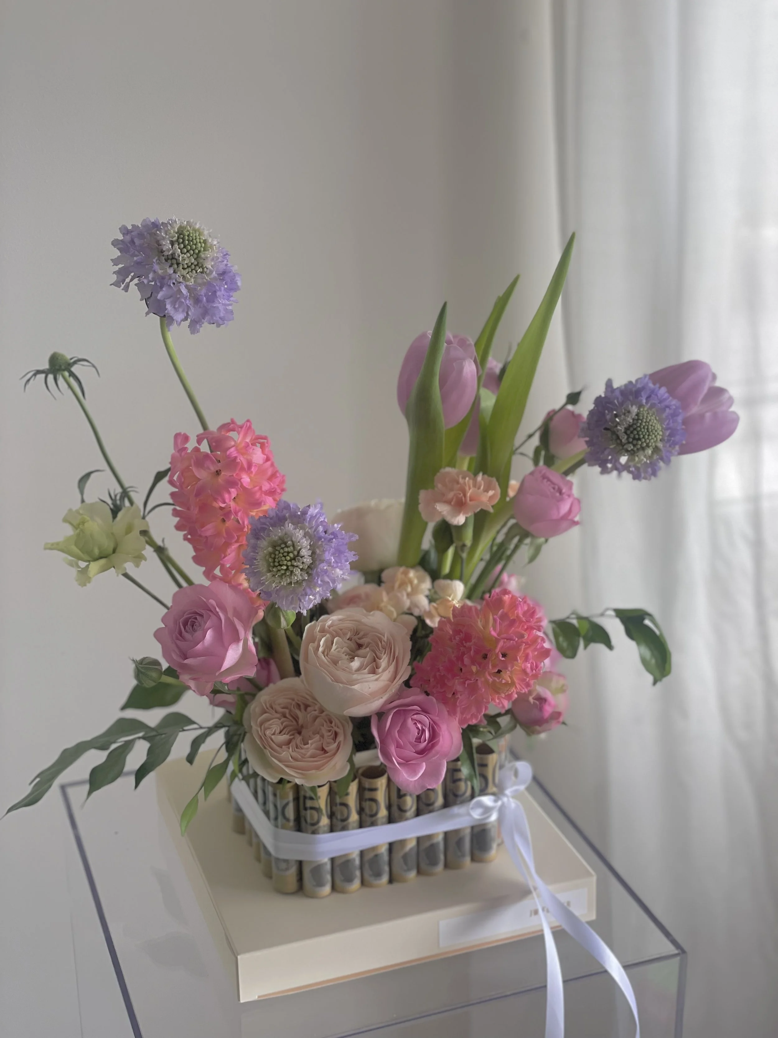 A floral arrangement with pink, purple, and cream flowers in a decorated container on a white stand, with a light-colored background and sheer curtains.