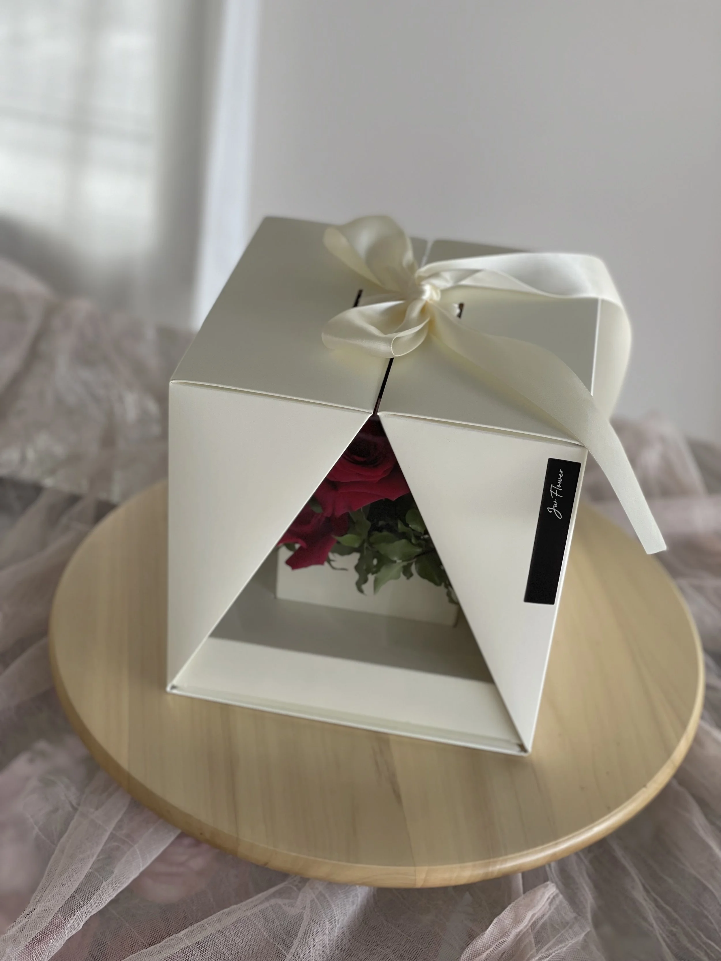 A white box with a black label that says 'Just Flavour' and a cream-colored satin ribbon tied in a bow on top, sitting on a round wooden surface. The box contains pink roses visible through a triangular opening.