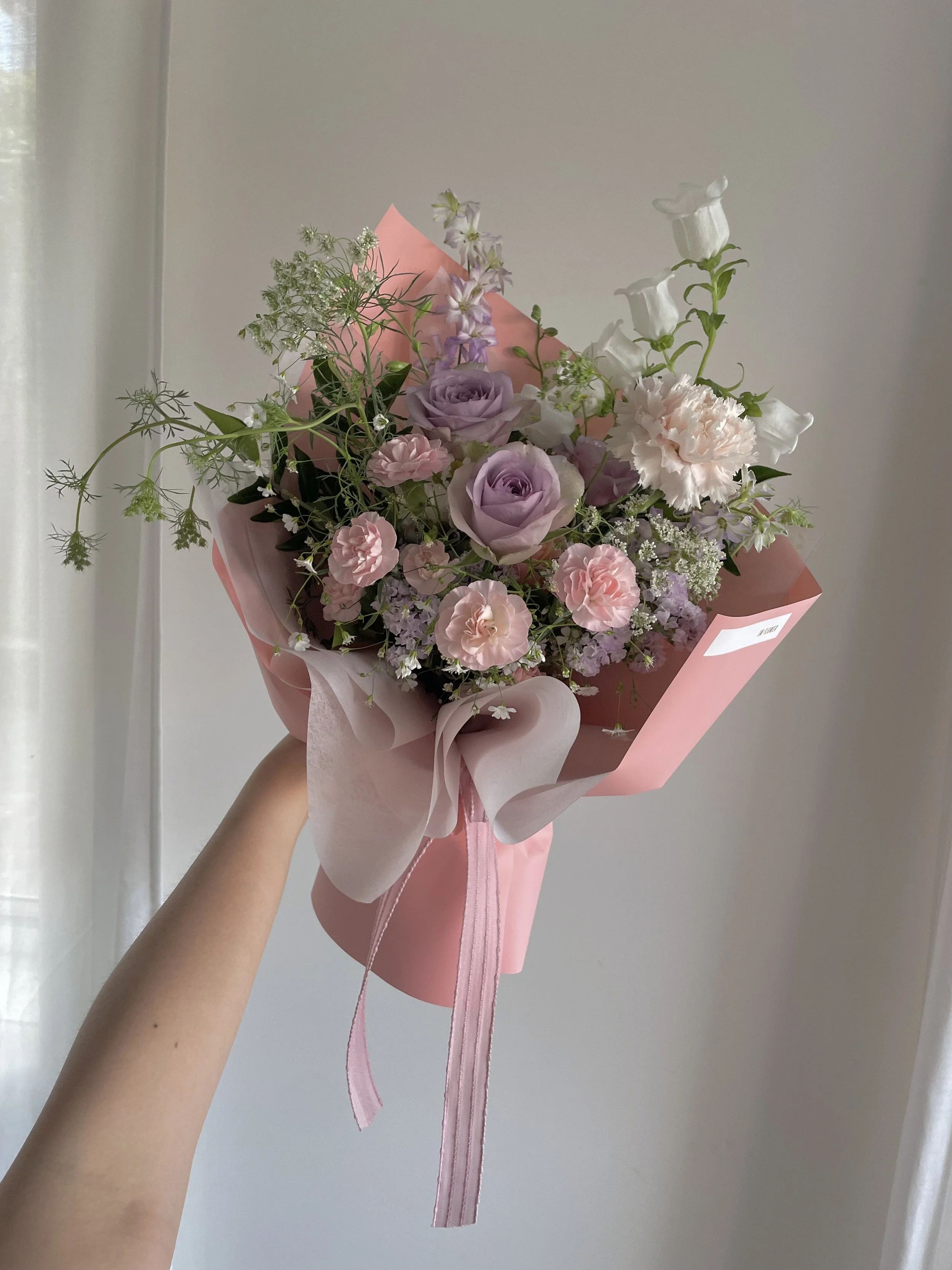 A hand holding a pink bouquet of flowers with roses, carnations, and greenery in pastel wrapping