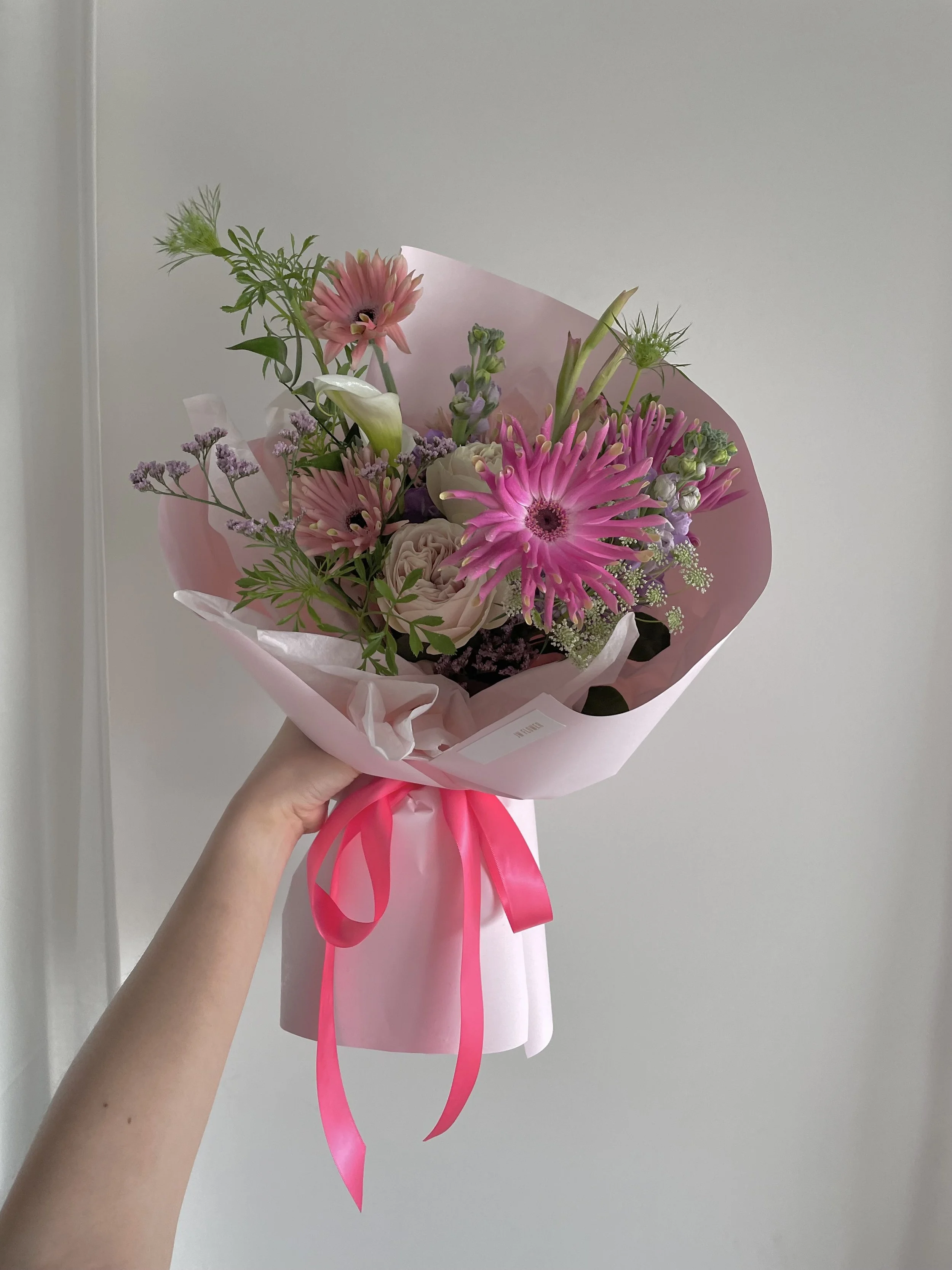 A hand holding a pink bouquet of various flowers including pink daisies, roses, and greenery, wrapped in pink paper with a pink ribbon against a plain gray wall.