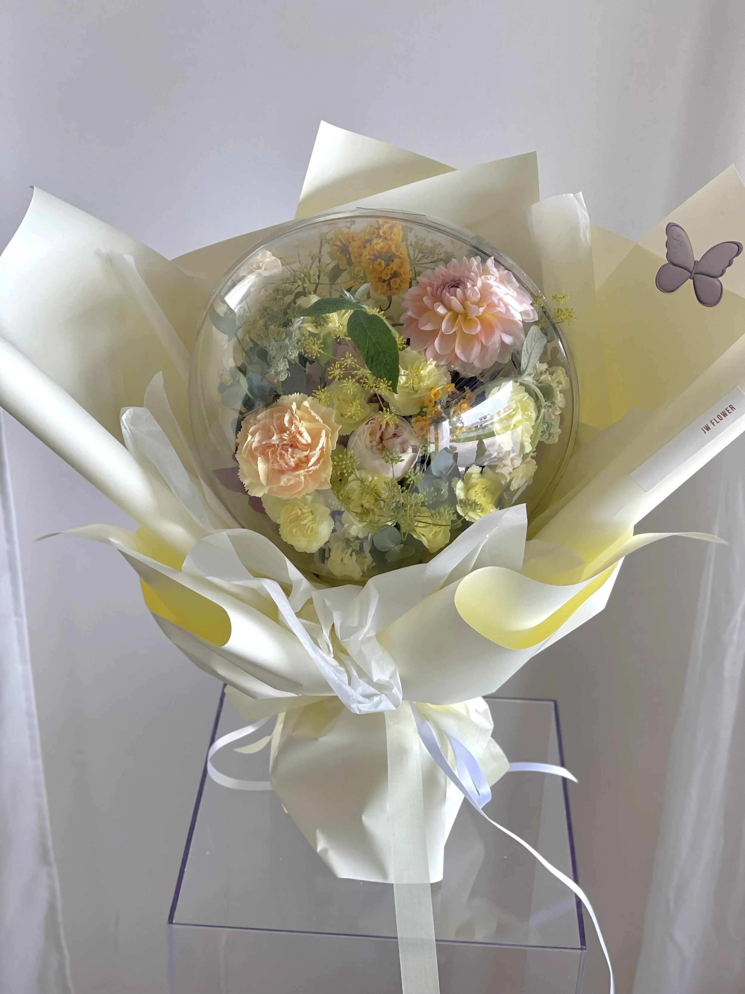 A bouquet of pastel-colored flowers, including pink and peach carnations, wrapped in white and yellow paper with a butterfly sticker, placed on a transparent surface.