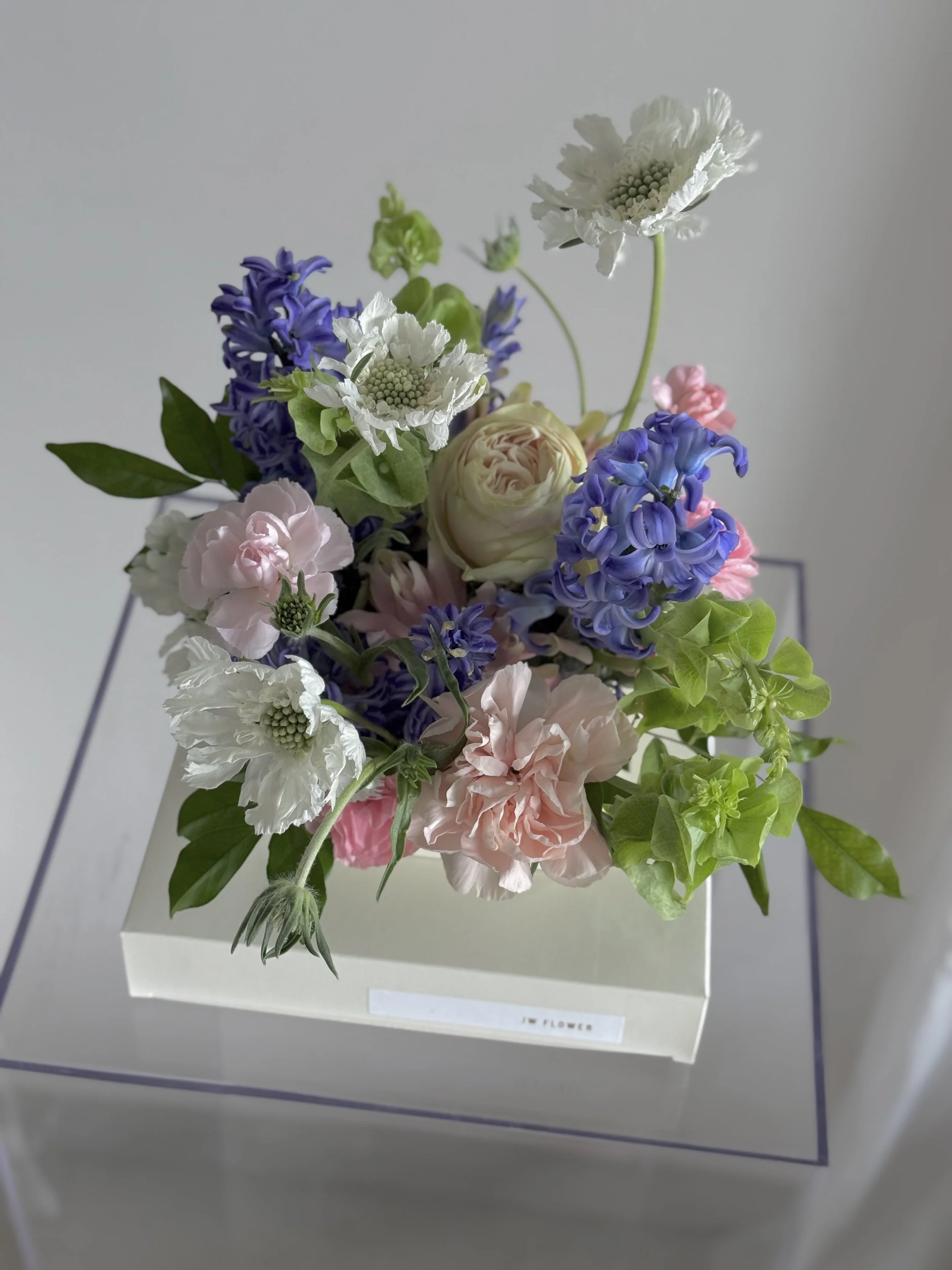 A bouquet of various pastel-colored flowers including white, pink, and purple blooms in a rectangular white box with a label that reads "JW FLOWER".