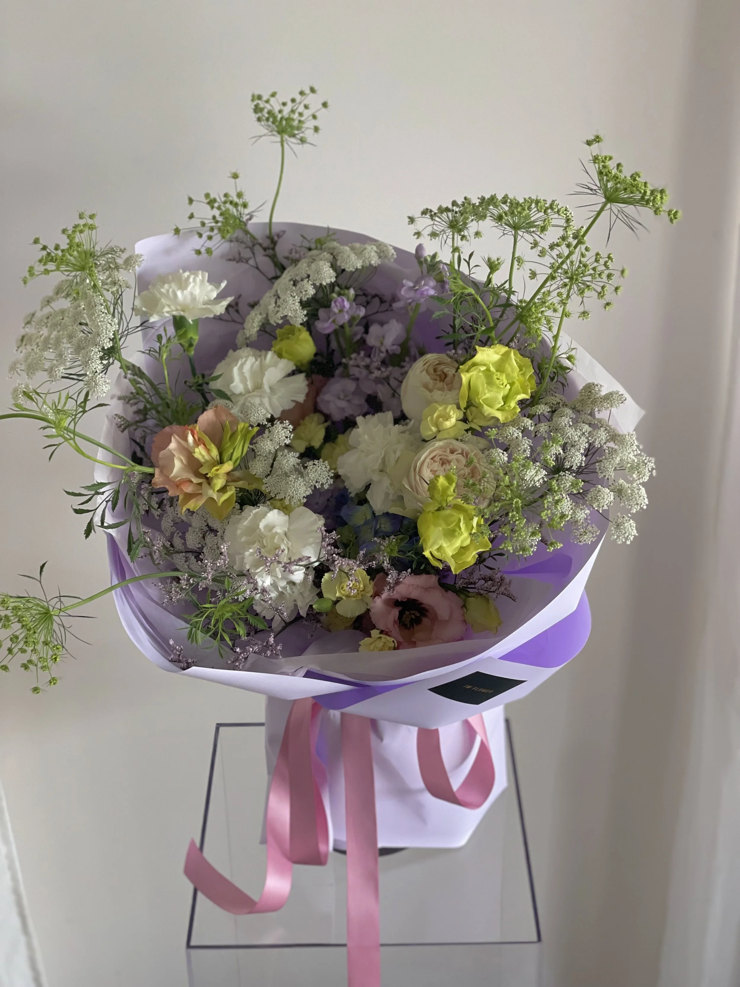 A bouquet of mixed flowers, including white, pink, and yellow blooms, wrapped in purple and white paper, with pink ribbons, displayed on a stand against a plain wall.
