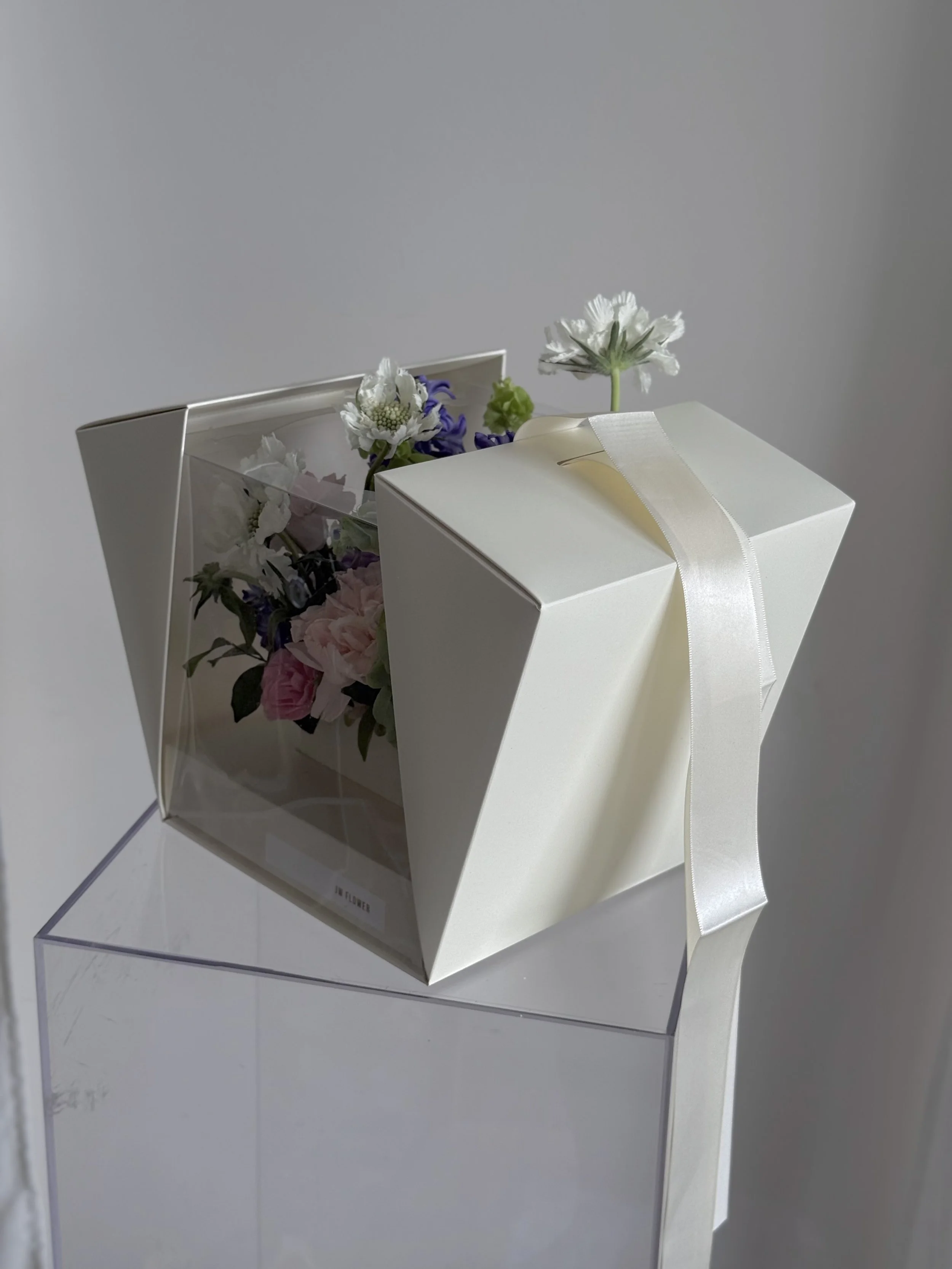 White gift box with a ribbon, decorated with flowers inside and out, placed on a clear stand against a plain background.