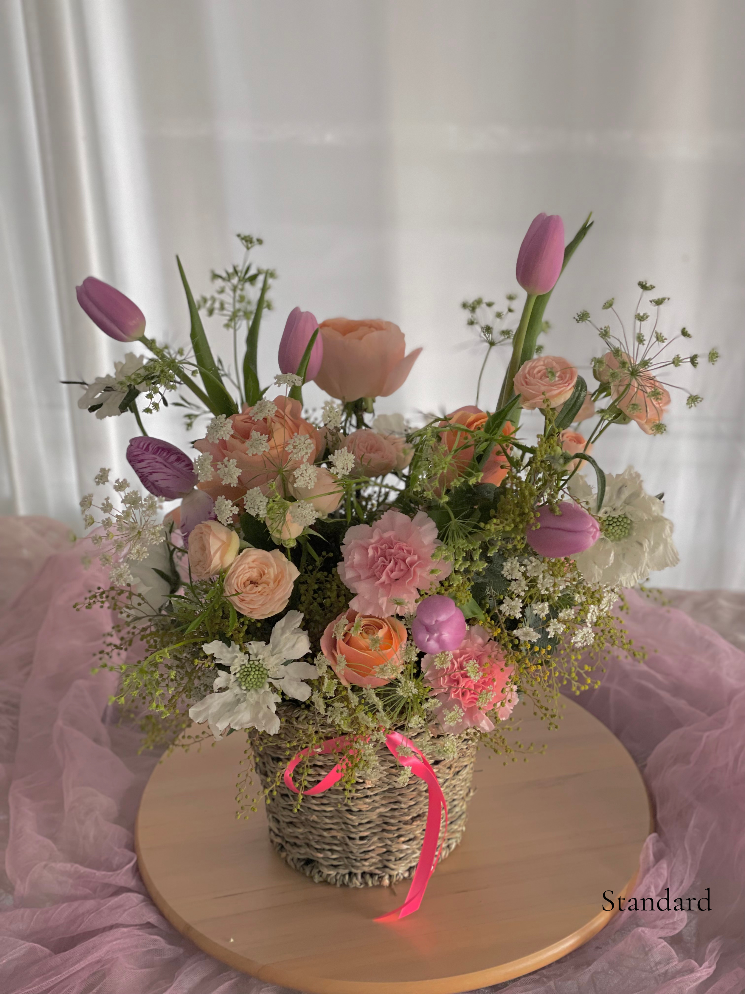 A floral arrangement with pink tulips, roses, carnations, and small white flowers in a woven basket with a pink ribbon, placed on a round wooden table with pink tulle fabric underneath.