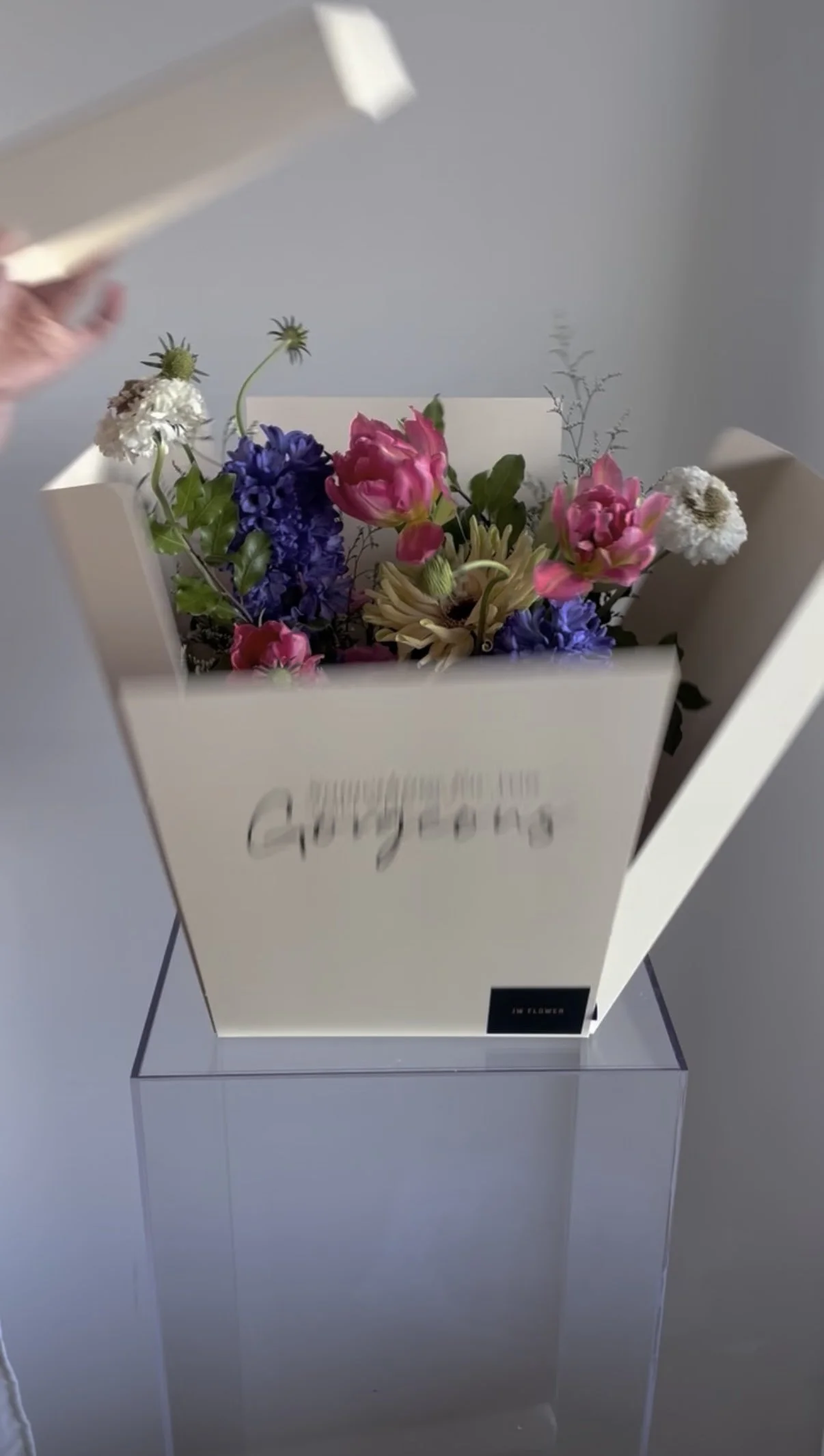 A white box with a floral arrangement of pink, purple, white, and cream flowers inside, placed on a clear display stand.