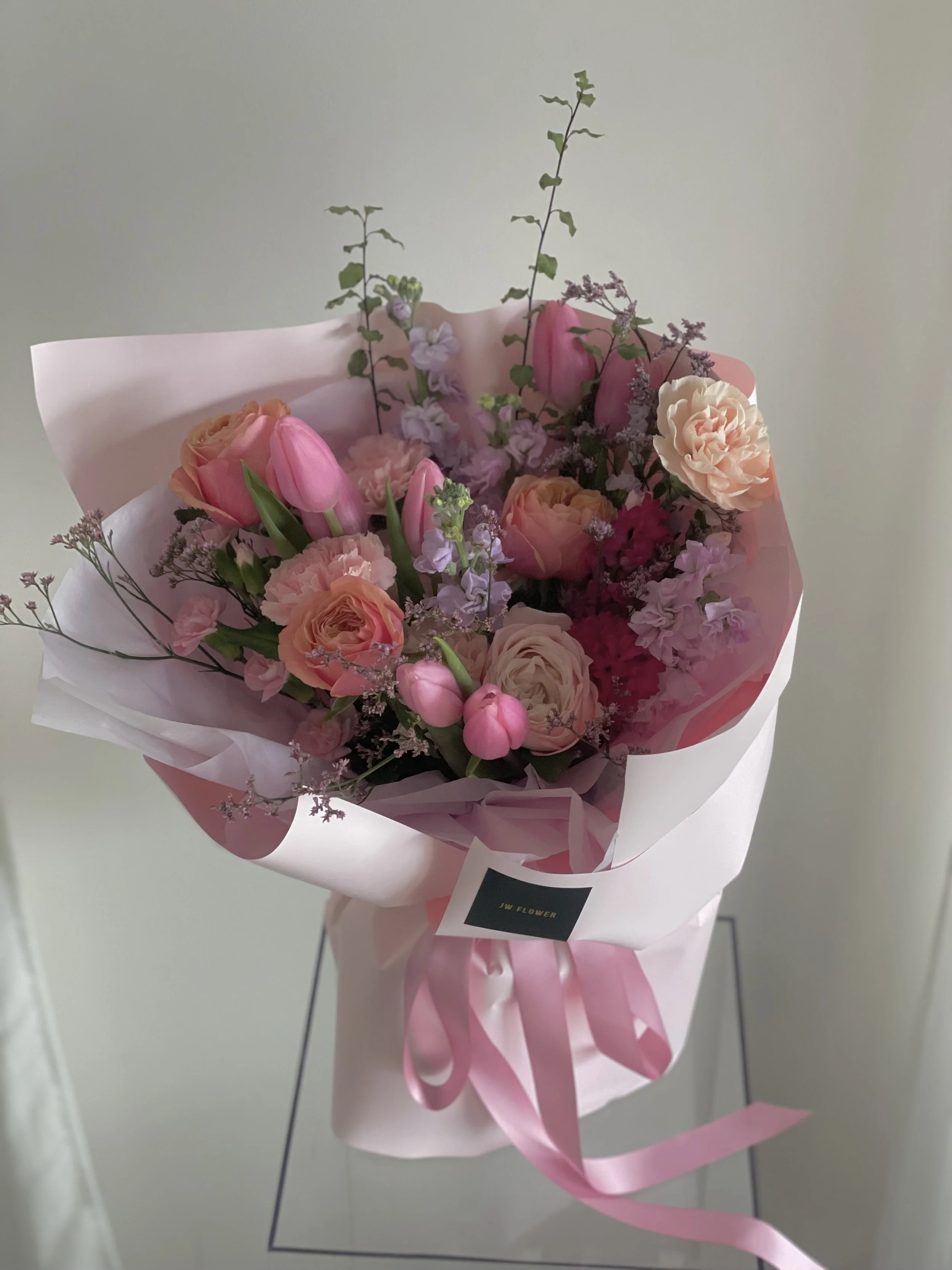 A bouquet of pink tulips, peach roses, and light purple stock flowers wrapped in pink and white paper with a pink ribbon, displayed on a stand against a plain wall.