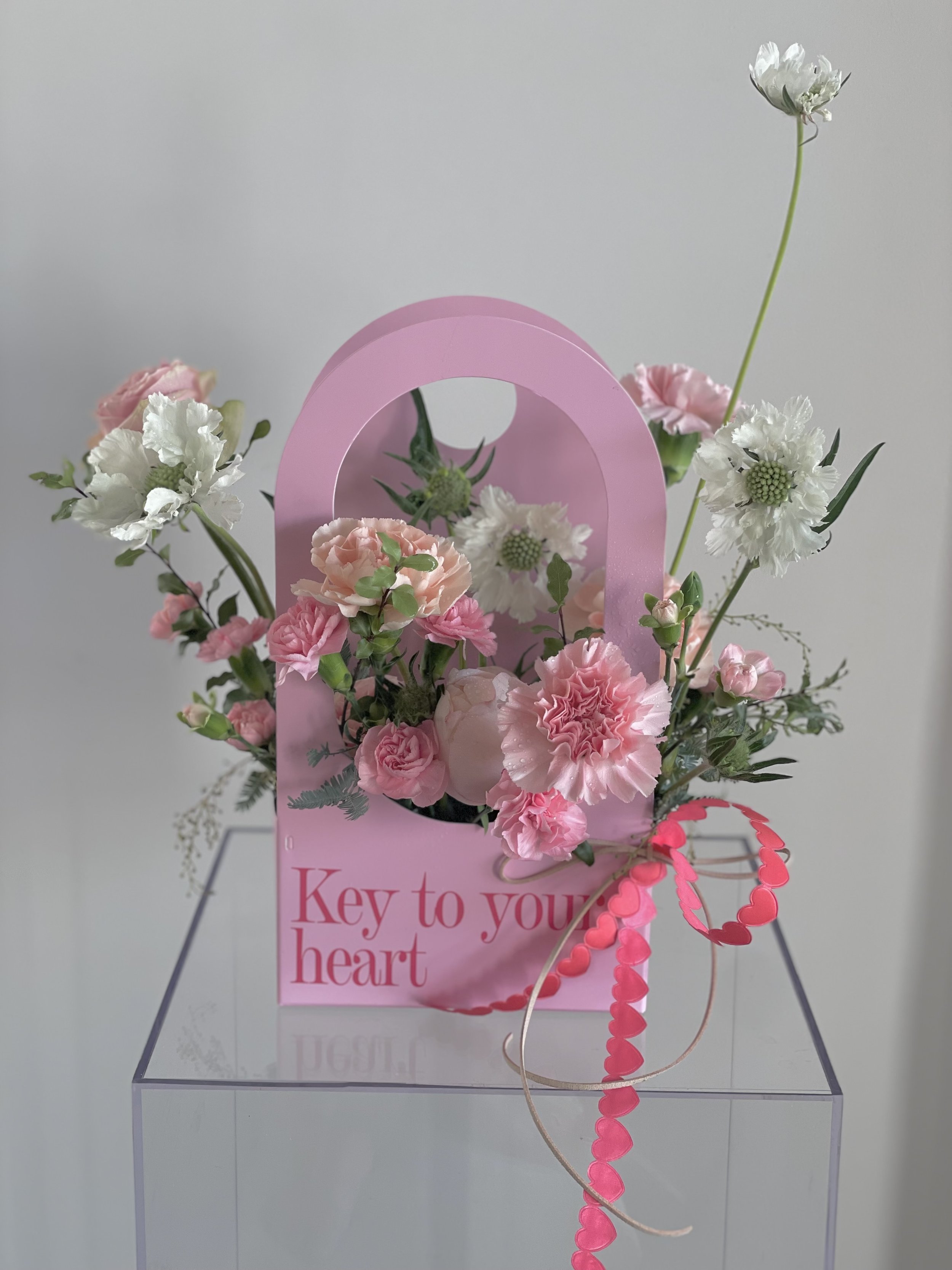 Pink floral arrangement in a pink decorative holder with the phrase "Key to your heart" written on it, placed on a clear stand against a plain background.