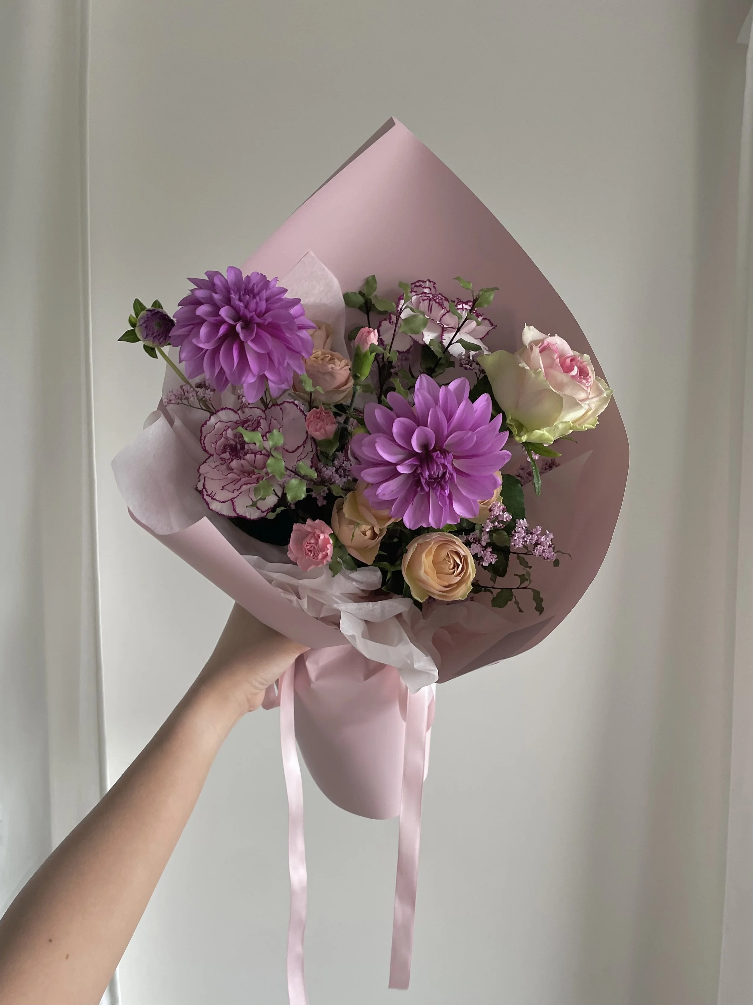 A hand holding a pink bouquet of flowers with purple dahlias, light pink roses, and greenery, wrapped in pink tissue paper.