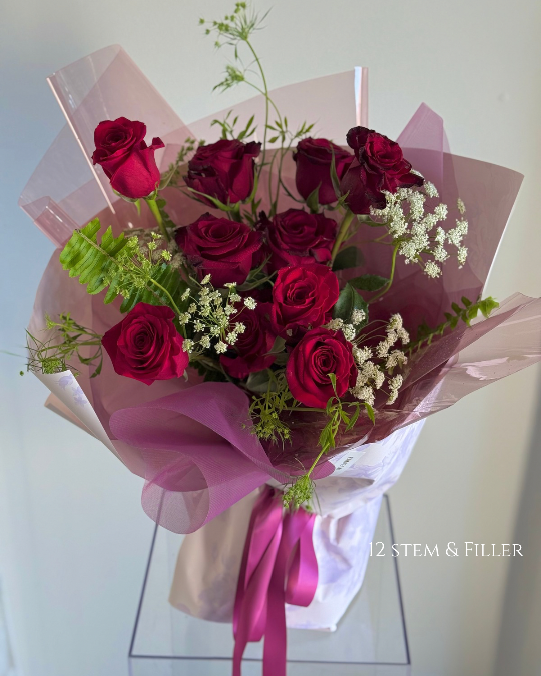 Bouquet of red roses with white baby's breath and green foliage, wrapped in pink and purple translucent paper with a pink ribbon