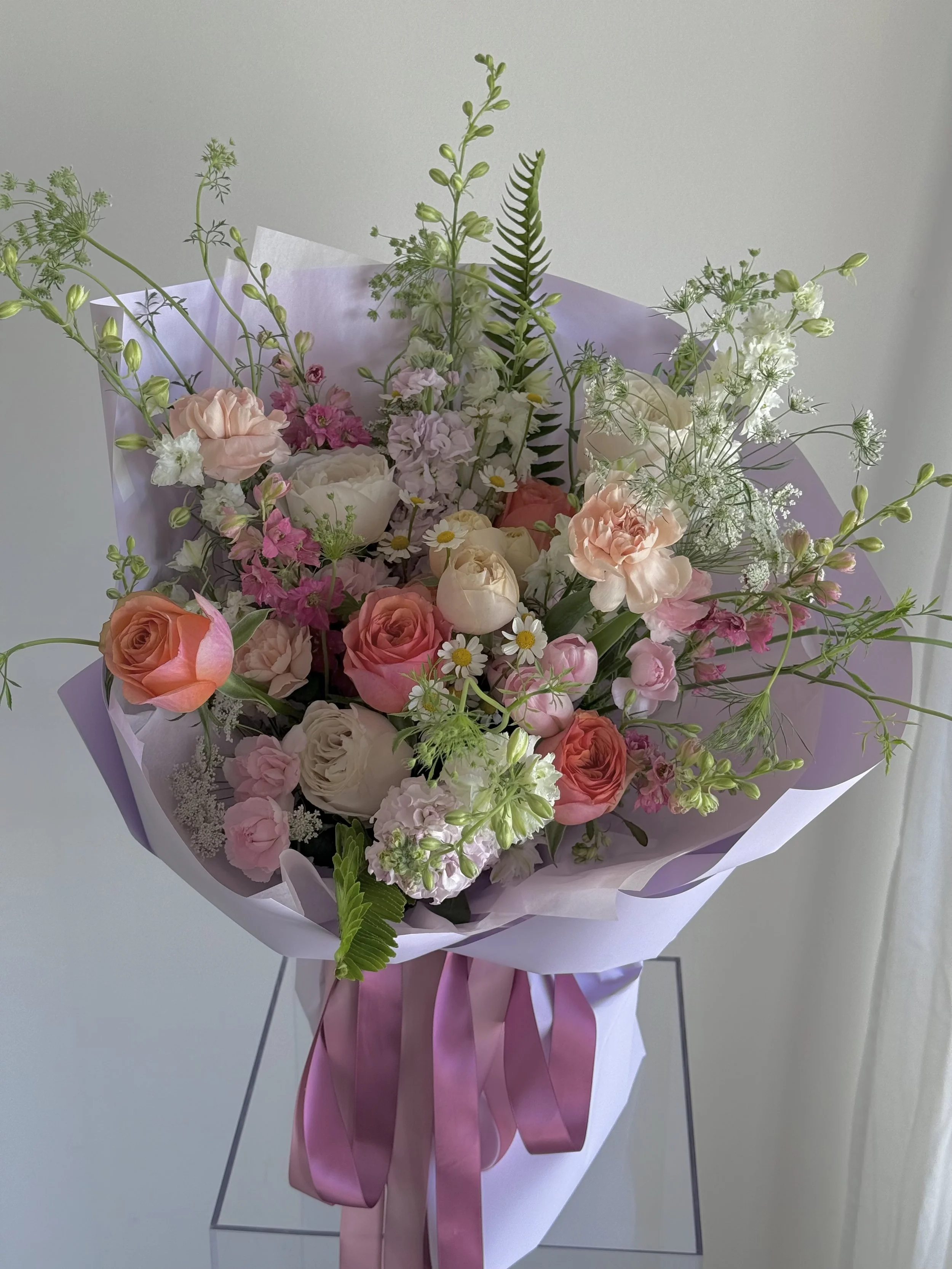 A bouquet of mixed flowers including roses, lisianthus, daisies, and greenery, wrapped in lilac paper with pink ribbons.