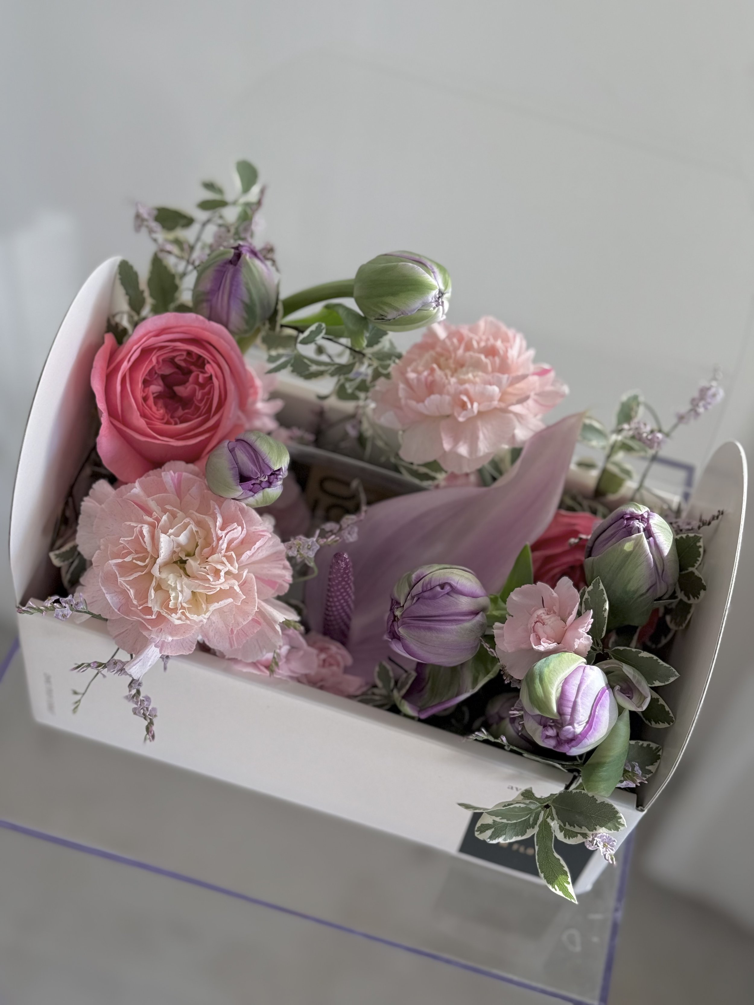 Arrangement of pink roses, carnations, and purple tulips in a white floral box.
