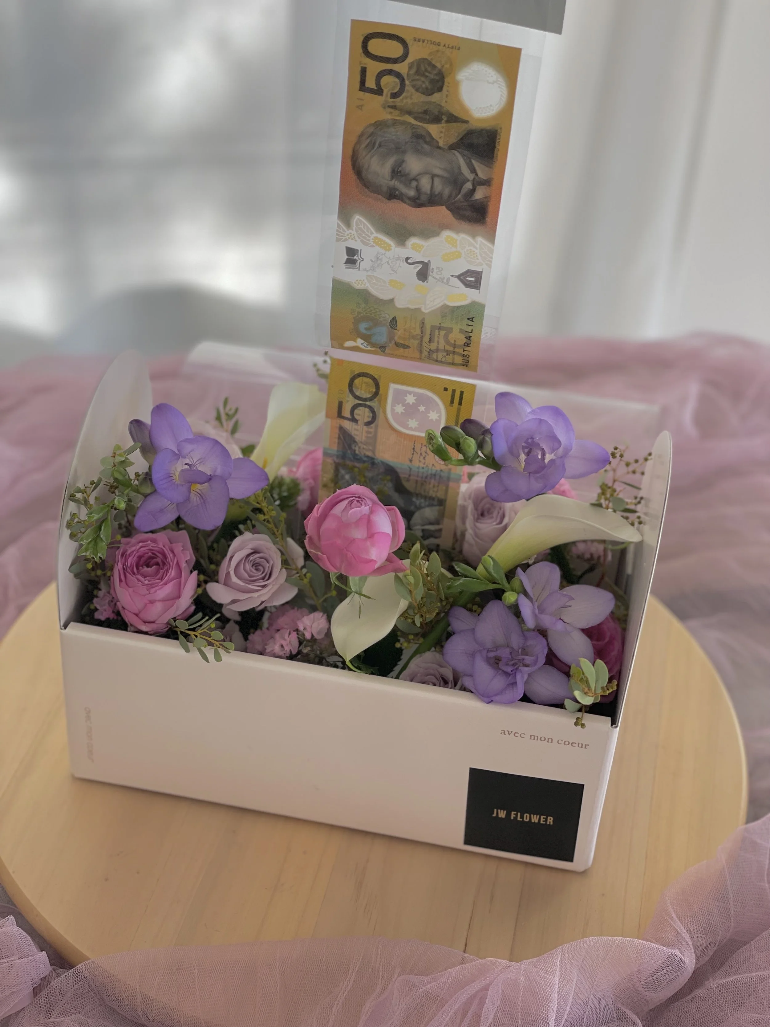 A flower arrangement with pink, purple, and white flowers, inside a white box labeled 'JW FLOWER,' with a 50 Australian dollar banknote standing upright among the flowers.