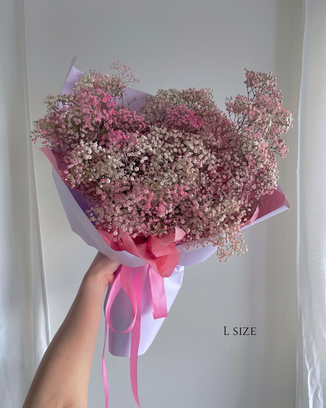 A large bouquet of pink and white baby's breath flowers wrapped in pink and purple paper with a pink ribbon. The bouquet is labeled 'L SIZE' and is held by a person's hand against a plain background.
