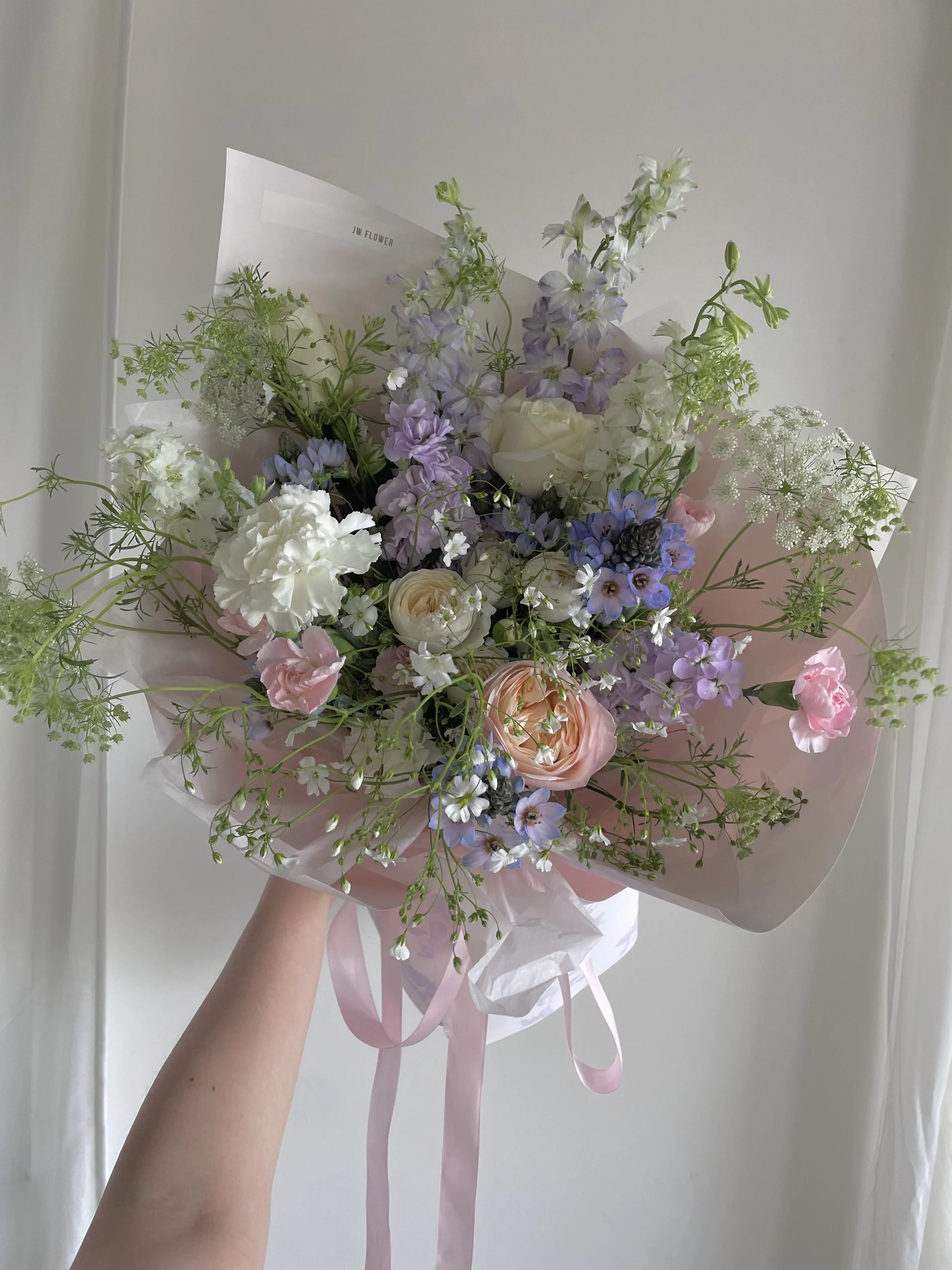 A bouquet of mixed flowers including roses, delphiniums, and other blooms in pastel colors, wrapped in pink and white paper with pink ribbons.