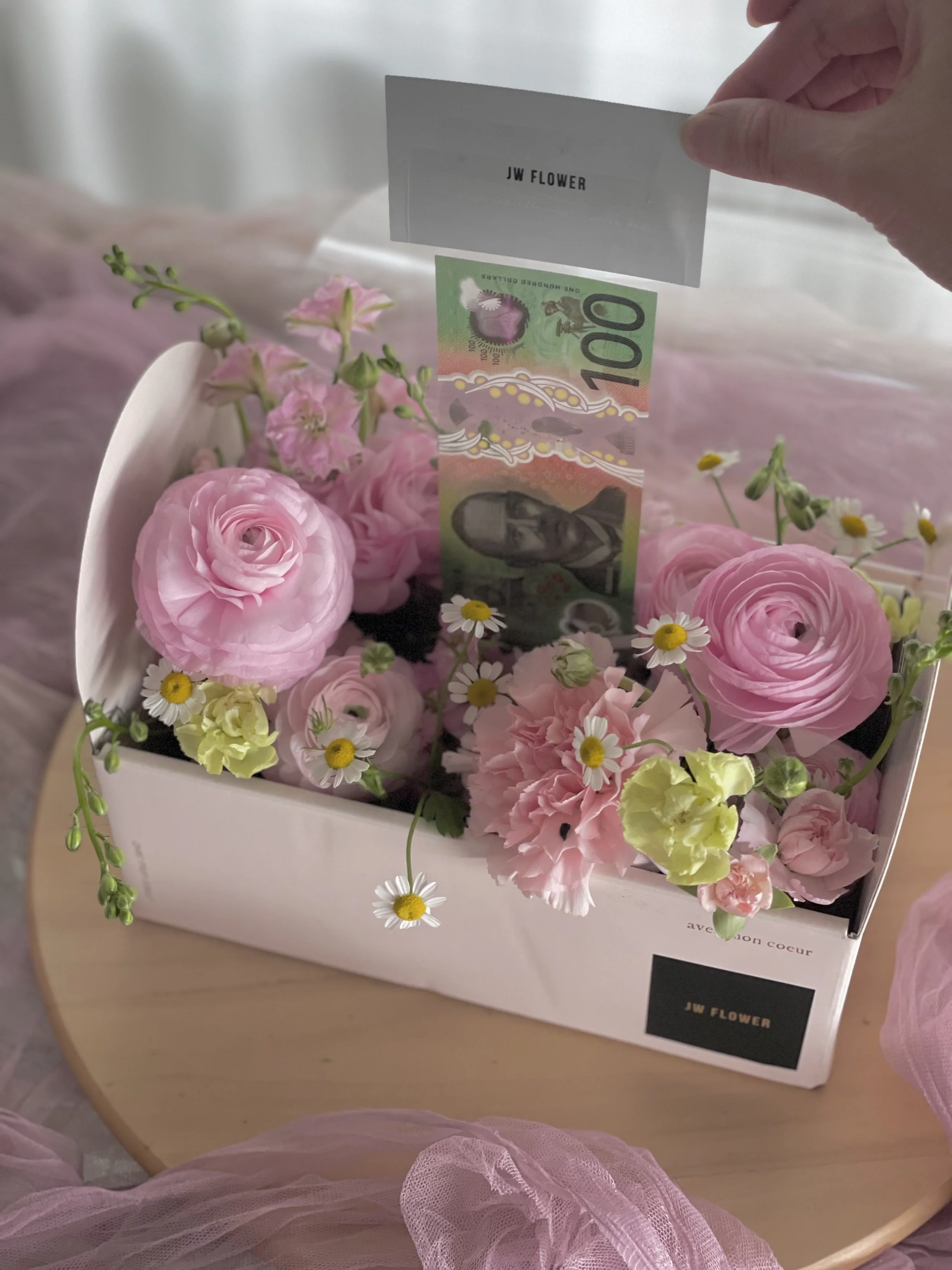 A white box filled with pink flowers and small daisies on a wooden table. A 100 Australian dollar bill is being inserted into the box with a note that says 'JW FLOWER'.