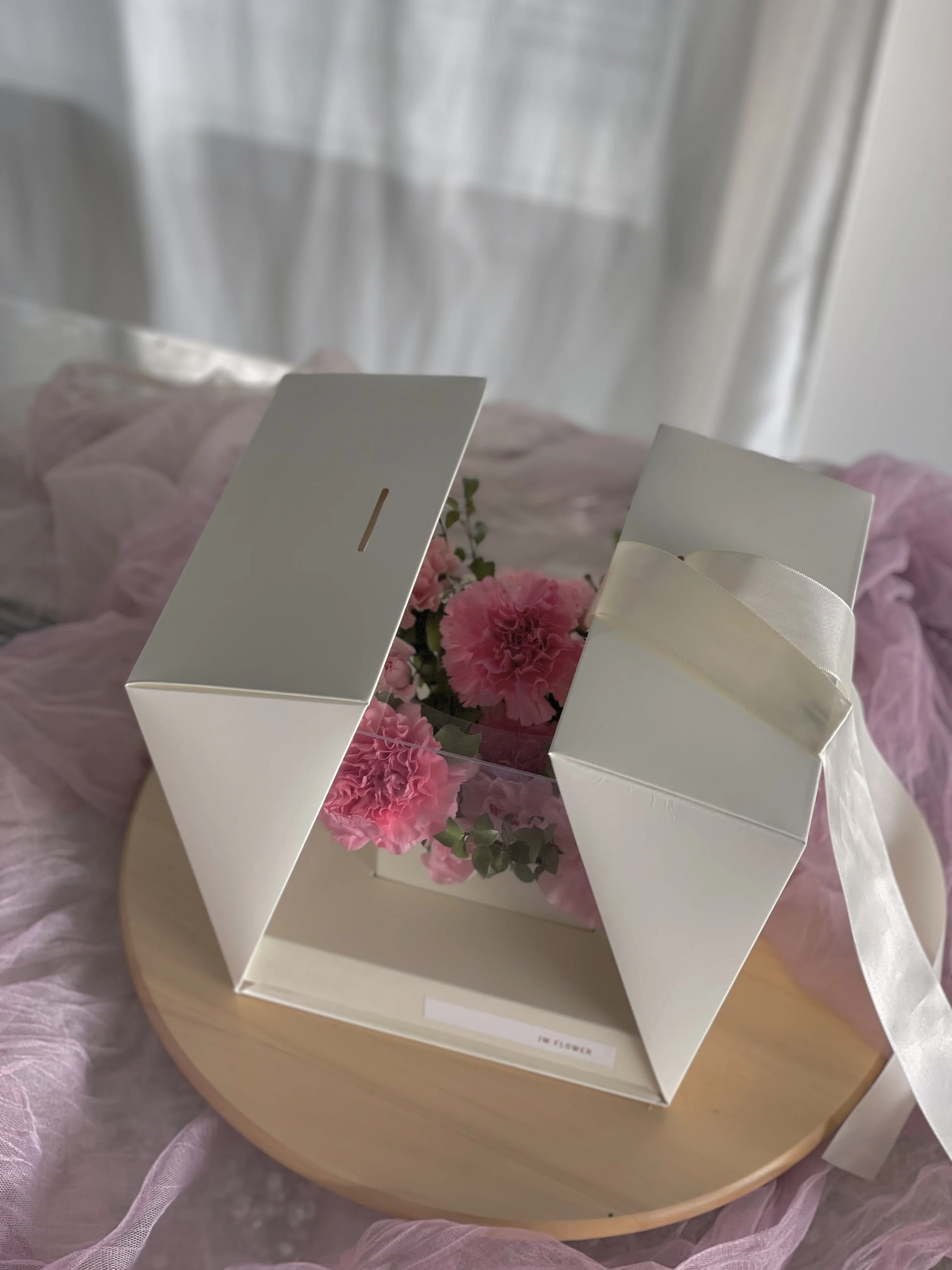 A gift box with handles, containing pink carnations and greenery, sitting on a round wooden tray, with pink fabric and curtains in the background.