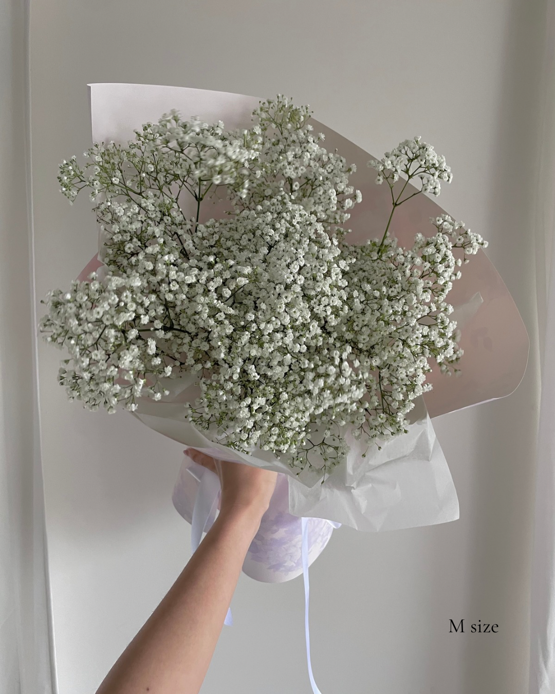 A hand holding a medium-sized bouquet of white baby's breath flowers wrapped in pink and white paper.