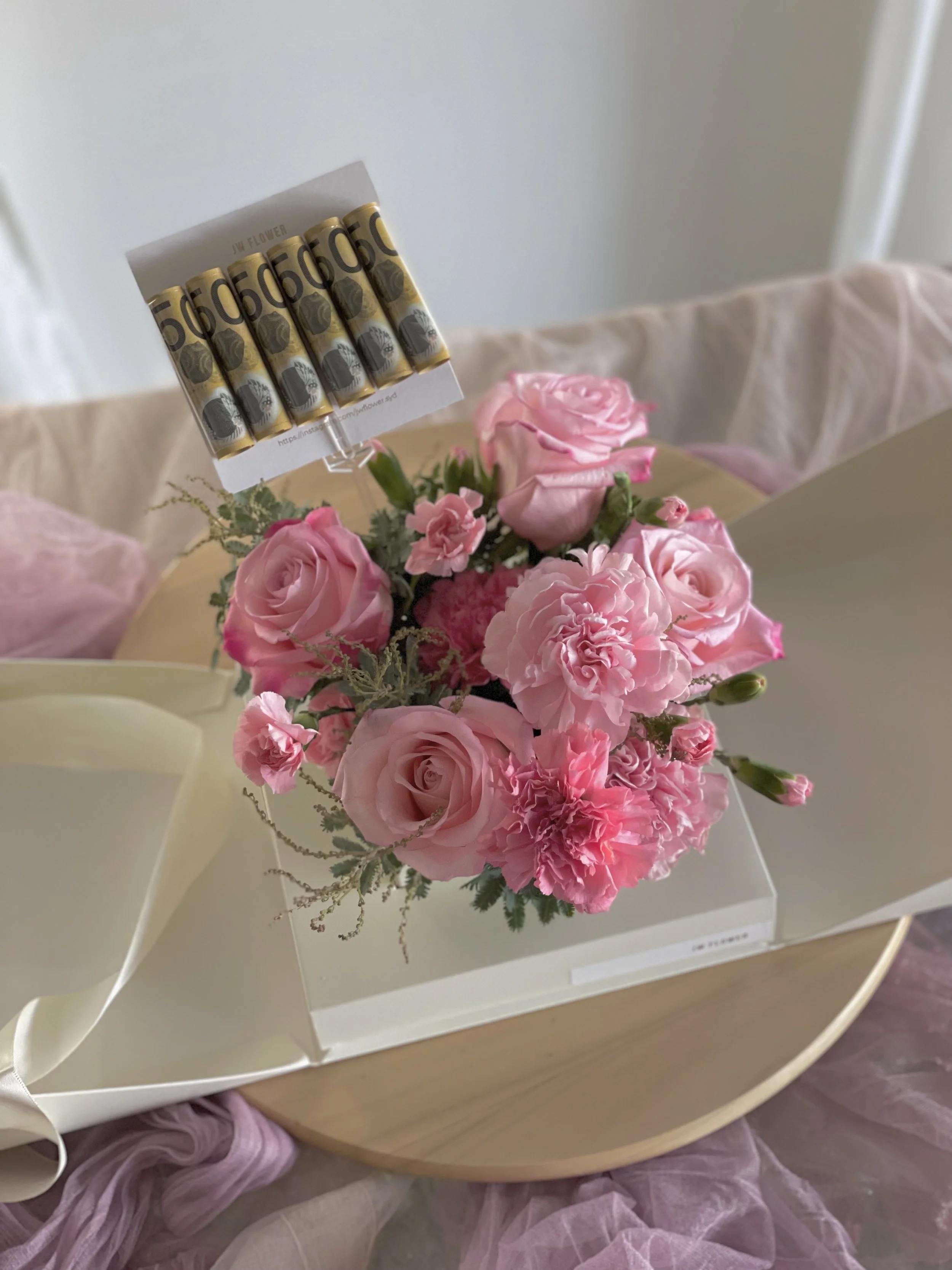 A bouquet of pink roses and carnations in a clear vial-shaped box with a white ribbon, placed on a wooden tray, with pink fabric draped nearby.