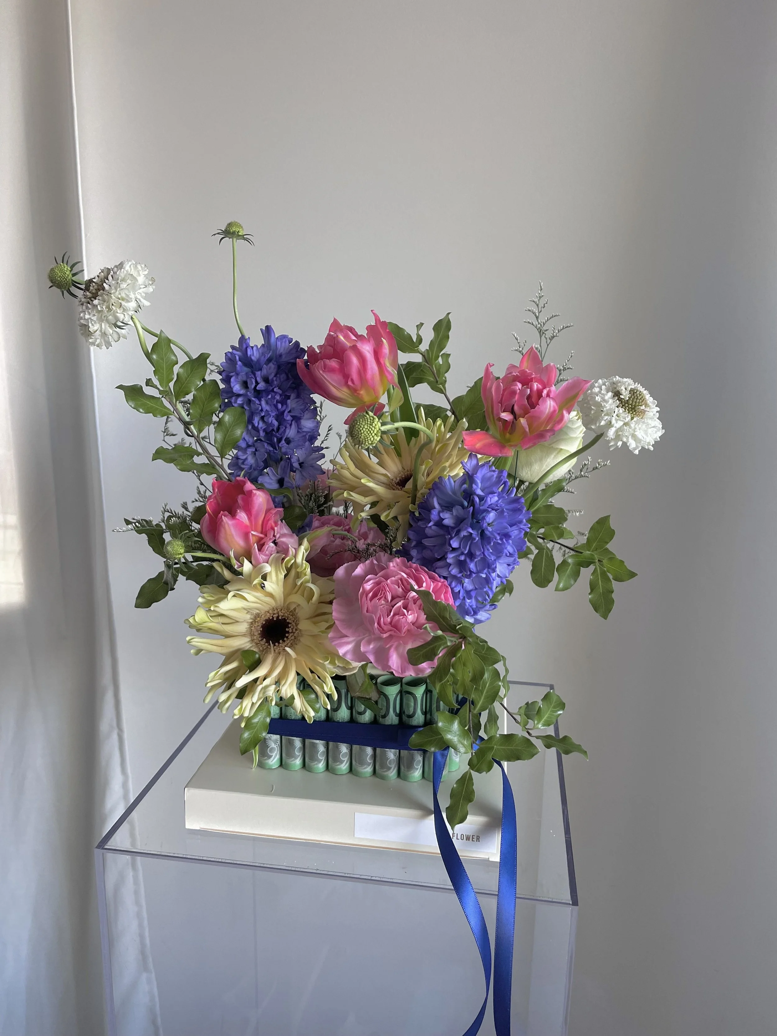 Colorful mixed flower bouquet with pink, white, purple, and cream flowers, arranged in a decorative container on a clear stand against a plain wall.