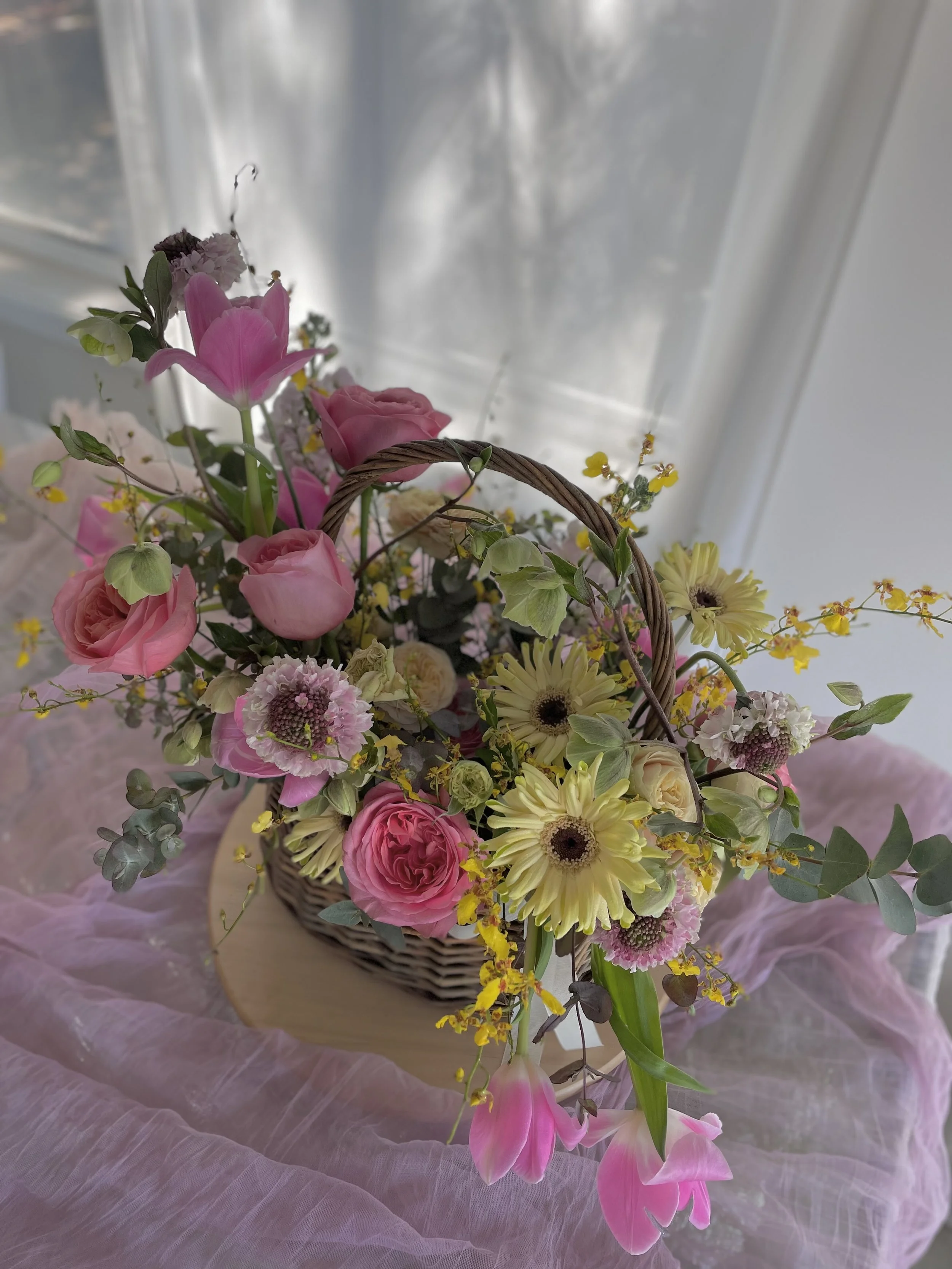 Pink, yellow, and white flower arrangement in a basket on pink fabric and a window in the background.