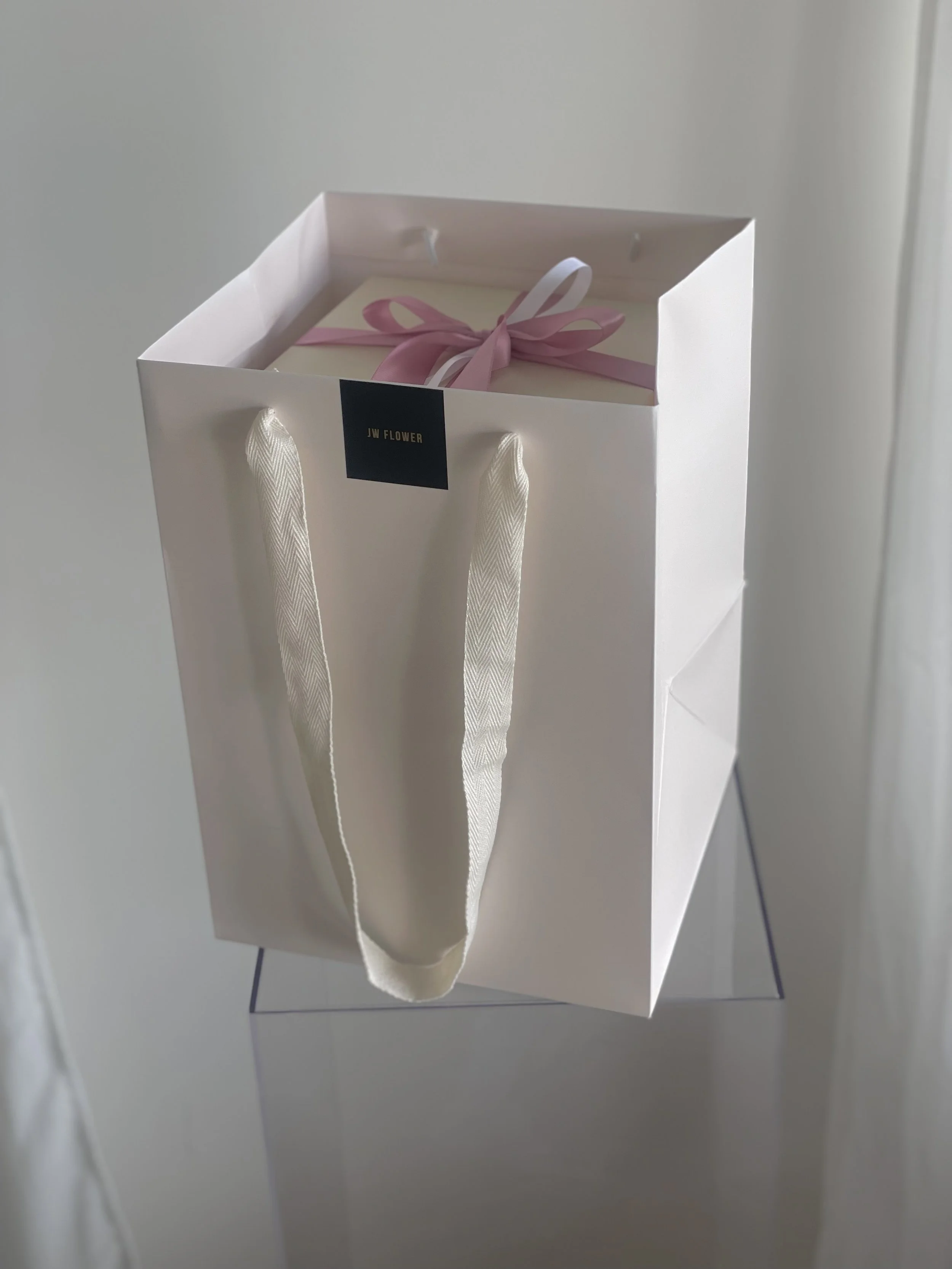 A white gift bag with satin handles and a small black label reading 'JW FLOWER' on the front. Inside, a pink and white box with a pink ribbon bow on top.