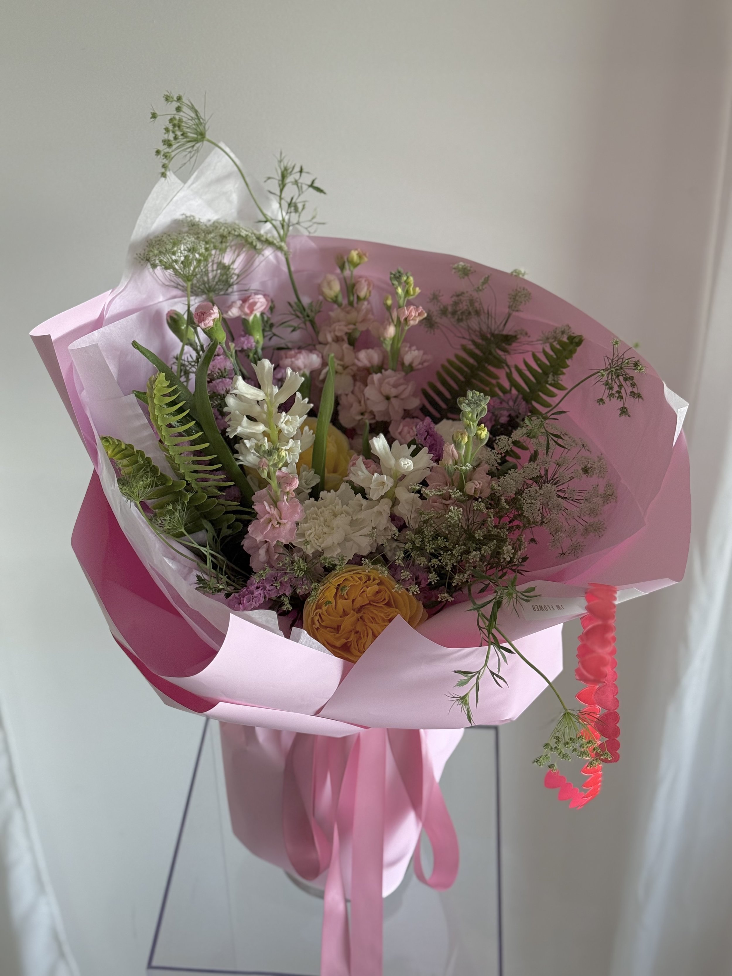 A bouquet of mixed flowers including pink, yellow, and white blossoms, wrapped in pink and white paper with ribbon, against a plain background.