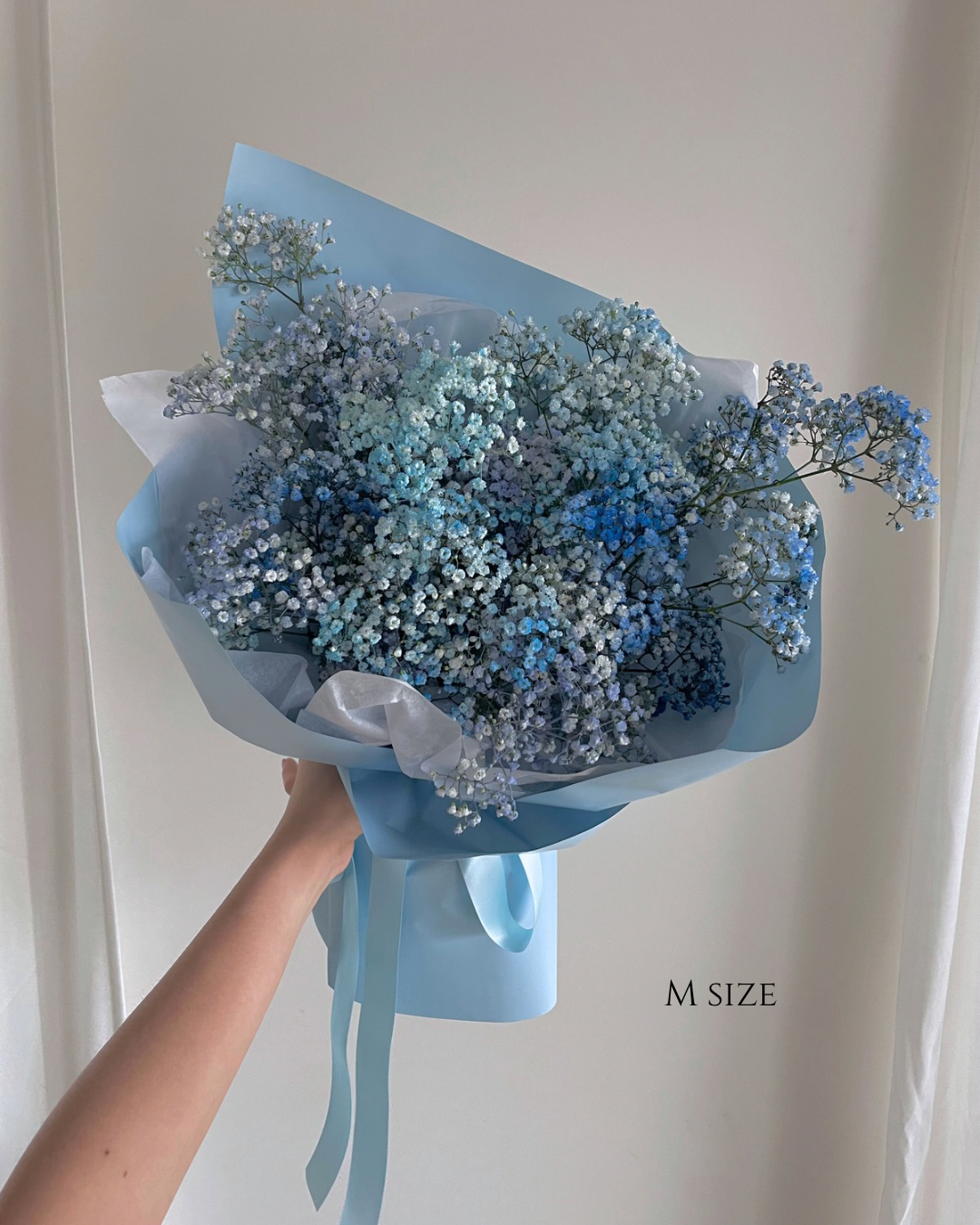 A hand holding a bouquet of baby's breath flowers wrapped in blue and white paper, with a blue ribbon, against a plain background.