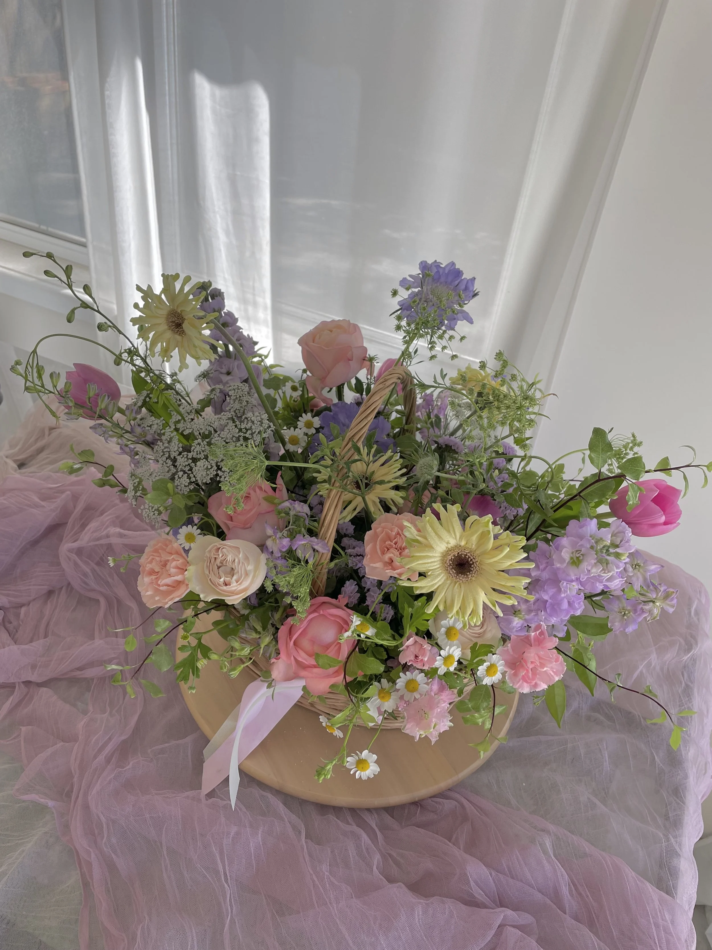 Arrangement of pink, purple, white, and yellow flowers in a wicker basket on pink tulle fabric.