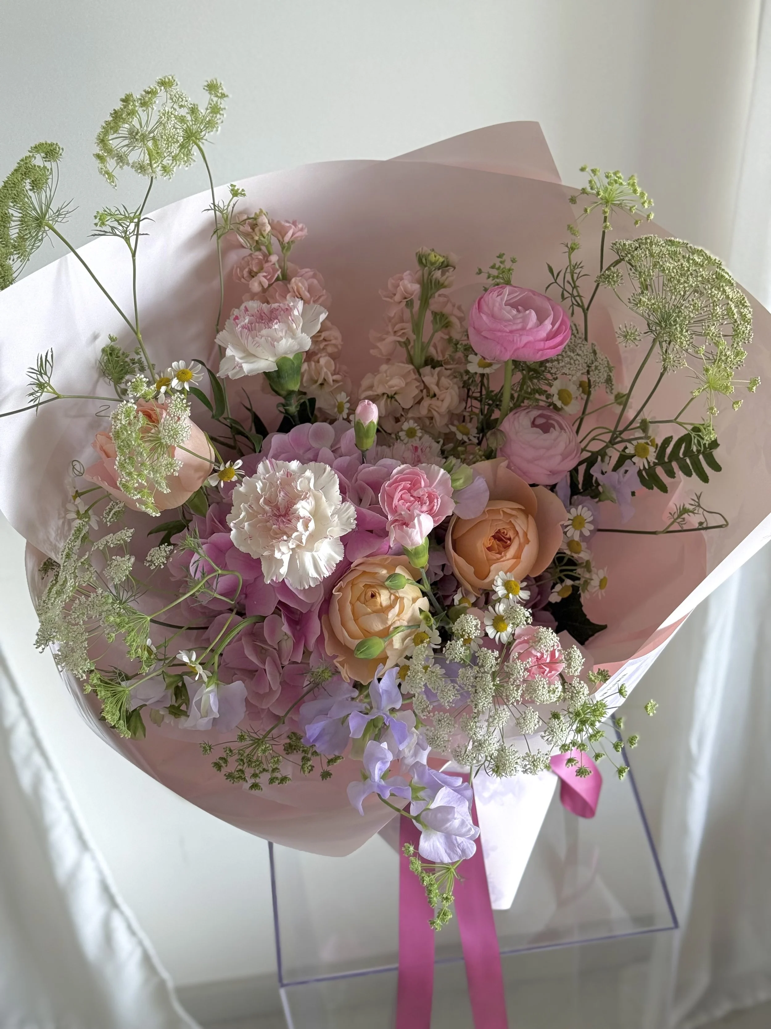A bouquet of pink, white, and peach flowers wrapped in light pink paper, tied with a pink ribbon.
