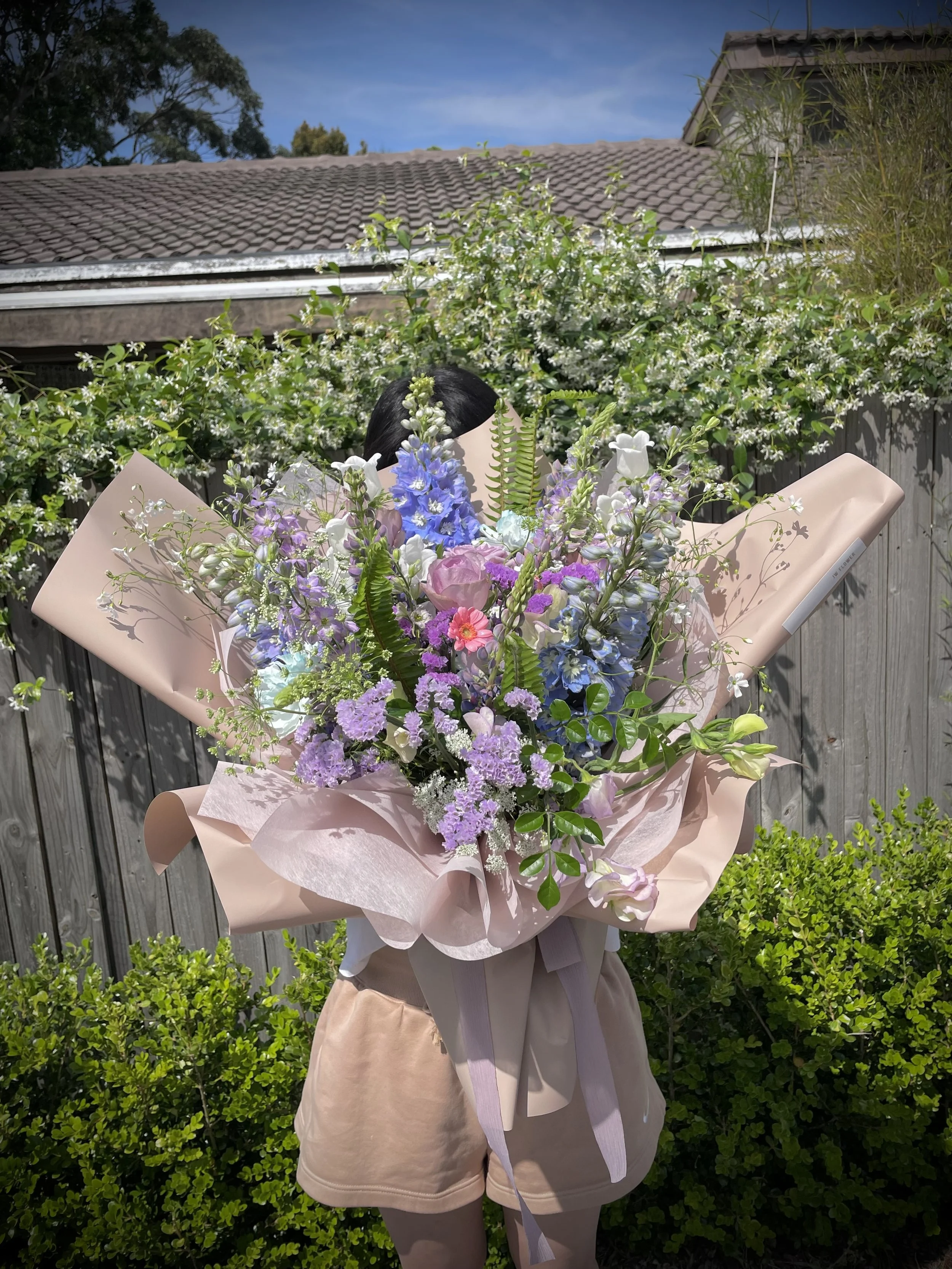 Person holding a large bouquet of mixed pastel-colored flowers outdoors in front of a wooden fence, with trees and a house roof in the background.