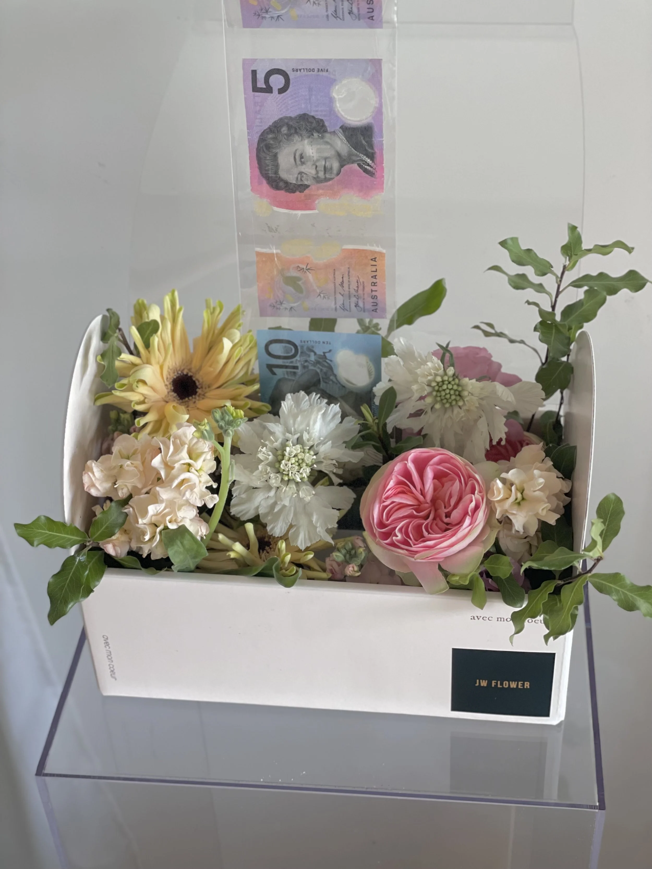 A floral arrangement with pink, white, and yellow flowers, green leaves, and issued Australian currency notes inside a white box labeled 'JW FLOWER.'