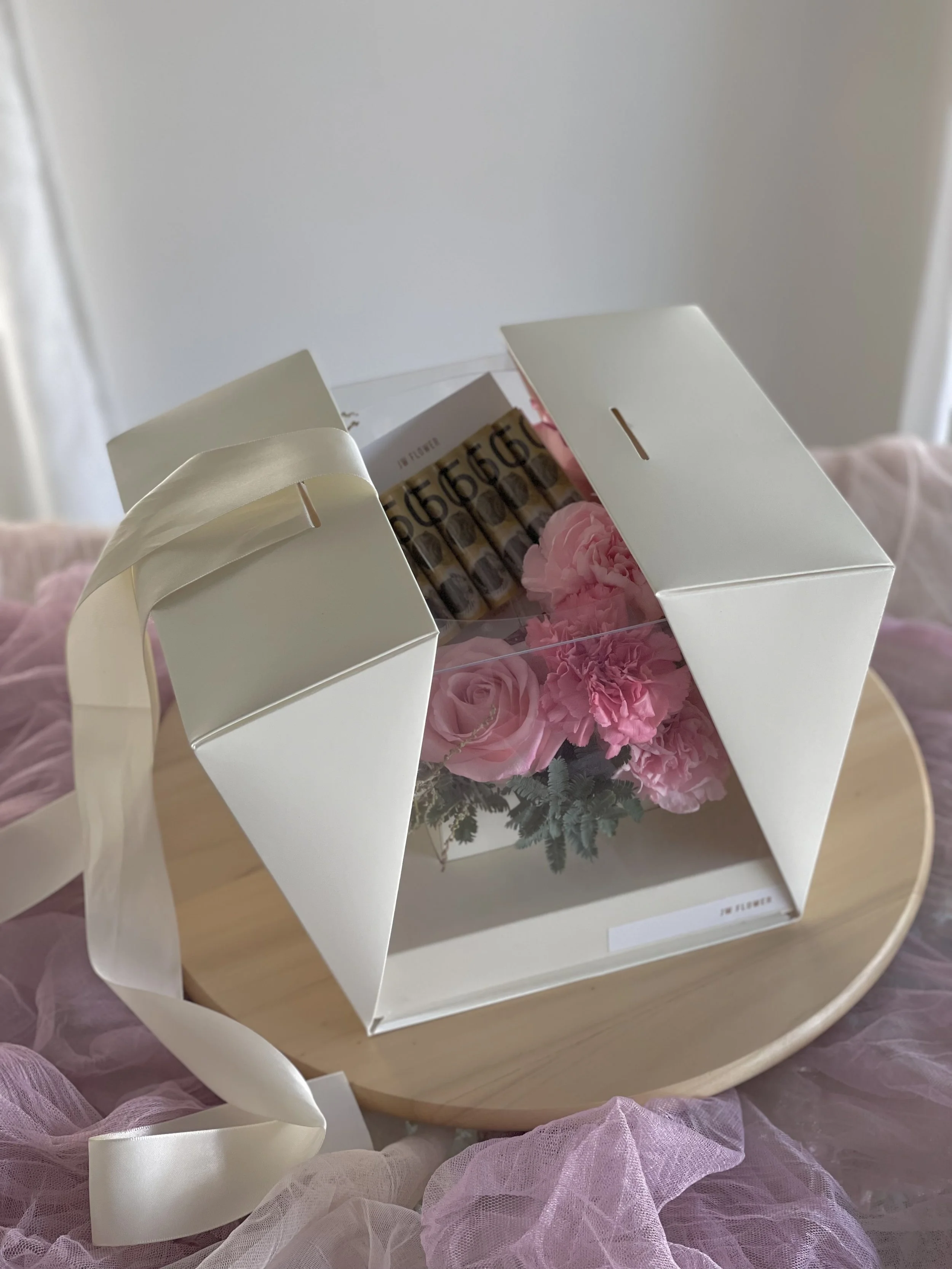 White box with a clear window on top, containing pink flowers and a set of small vials or bottles, placed on a round wooden surface with pink fabric around it.