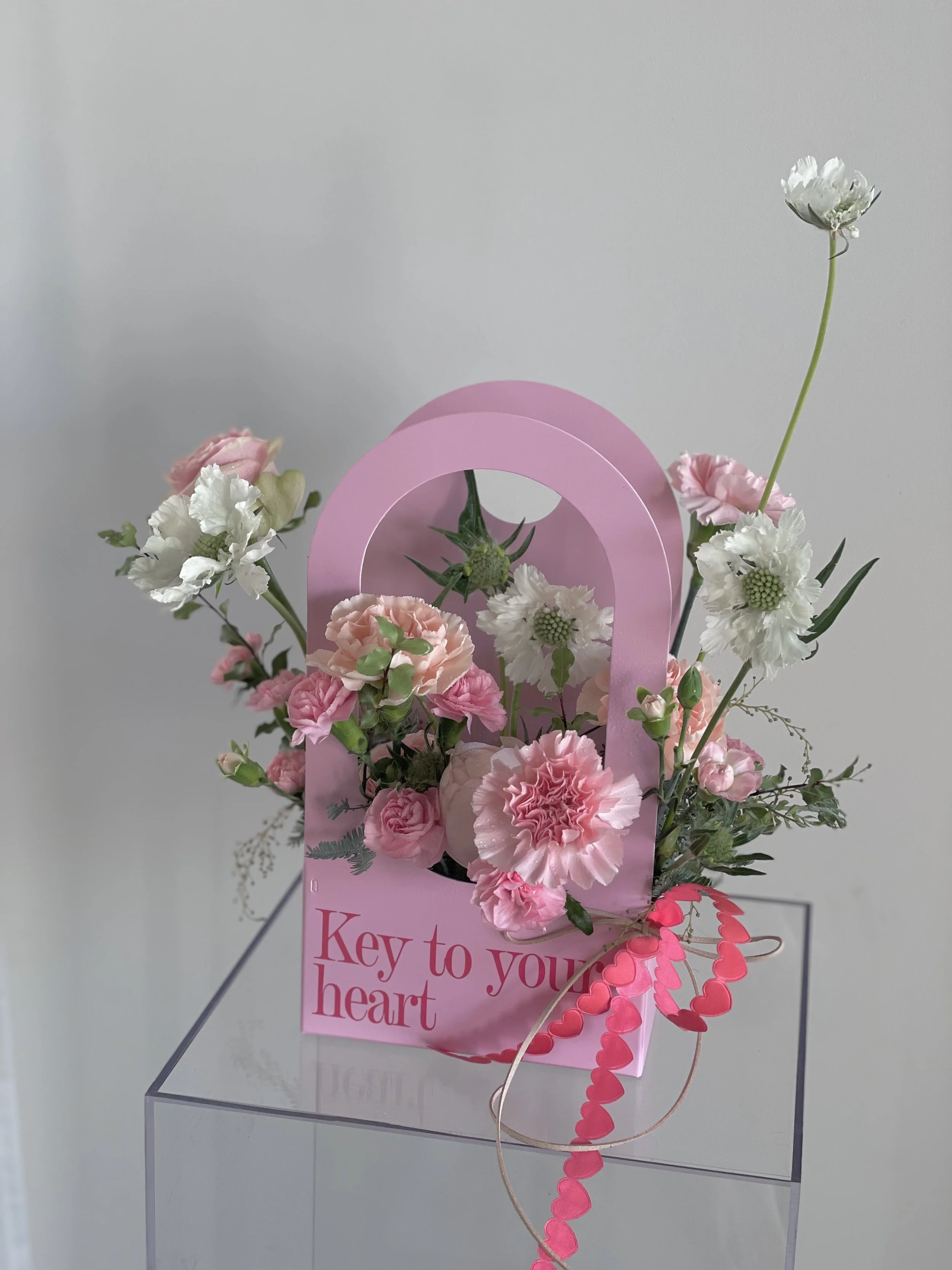 Pink floral arrangement inside a pink bag with a handle, with the words "Key to your heart" written on it. The arrangement includes pink and white flowers, some with ruffled petals, and green foliage. The bag and flowers are placed on a clear, box-shaped surface against a plain background.