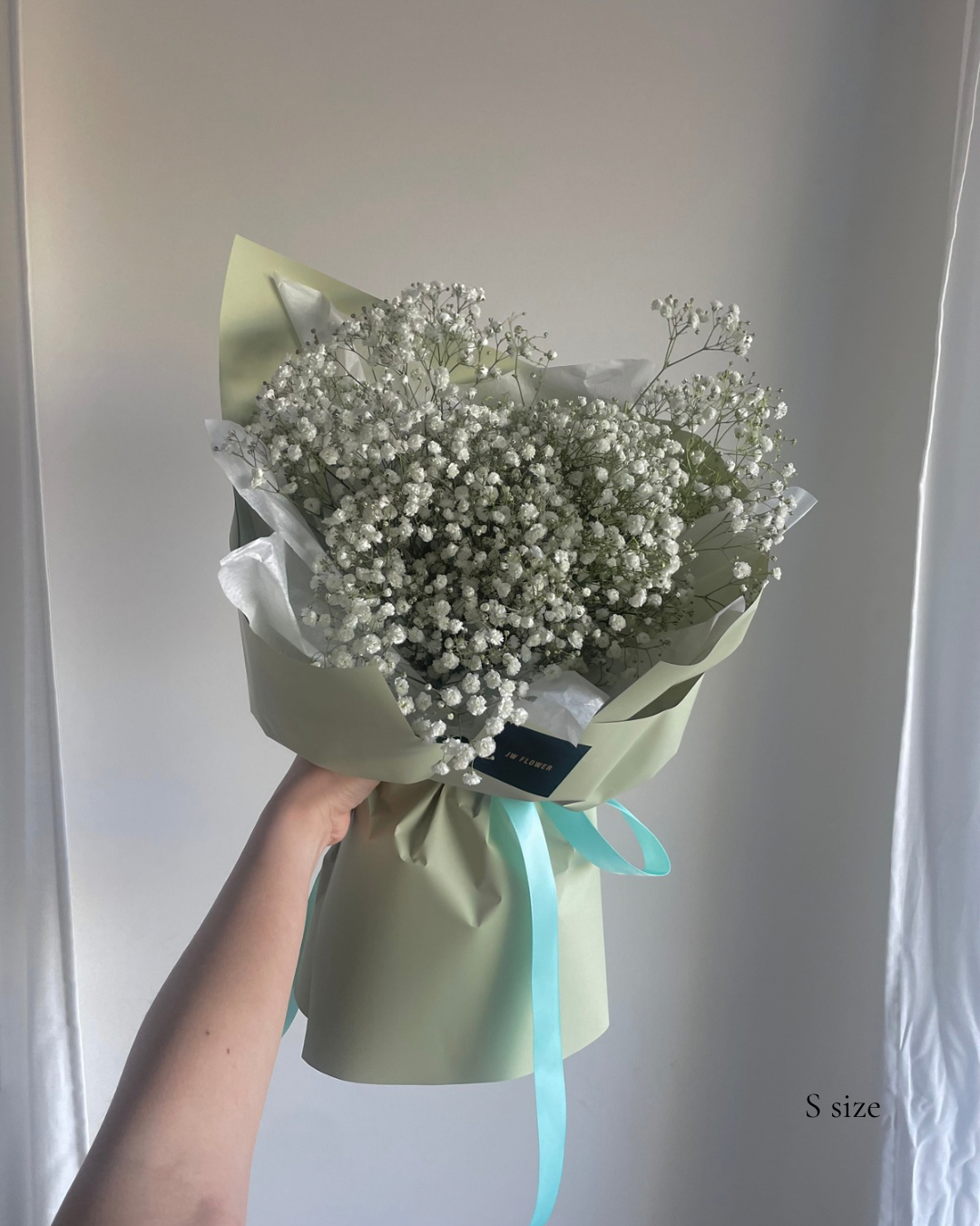 Hand holding a small bouquet of white baby's breath flowers wrapped in pale green paper with a light blue ribbon, against a plain background.