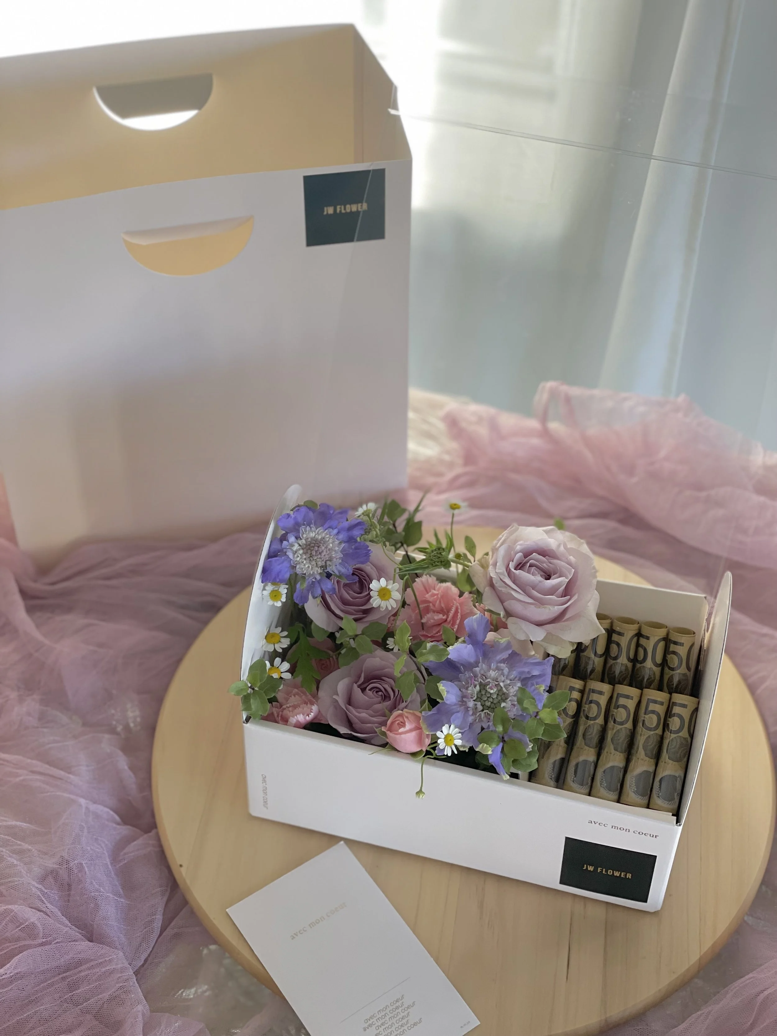 A white box filled with pastel purple roses, pink flowers, small white daisies, and purple flowers, alongside rolled banknotes, is placed on a round wooden table with pink fabric draped around it. In the background, there is a white shopping bag with a black label that says 'JW FLOWER'.