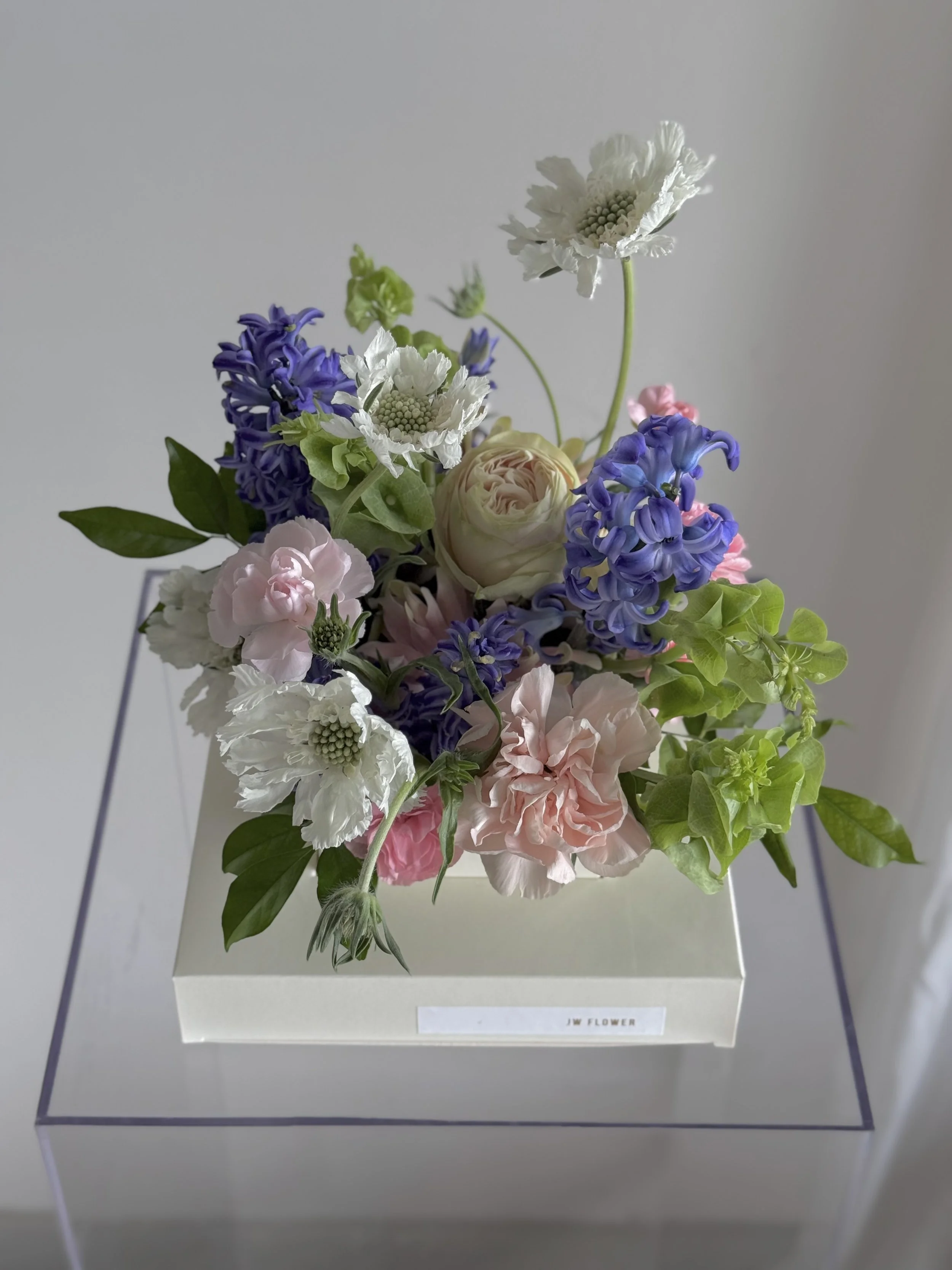 A colorful bouquet of mixed flowers in a white box, displayed on a transparent stand.