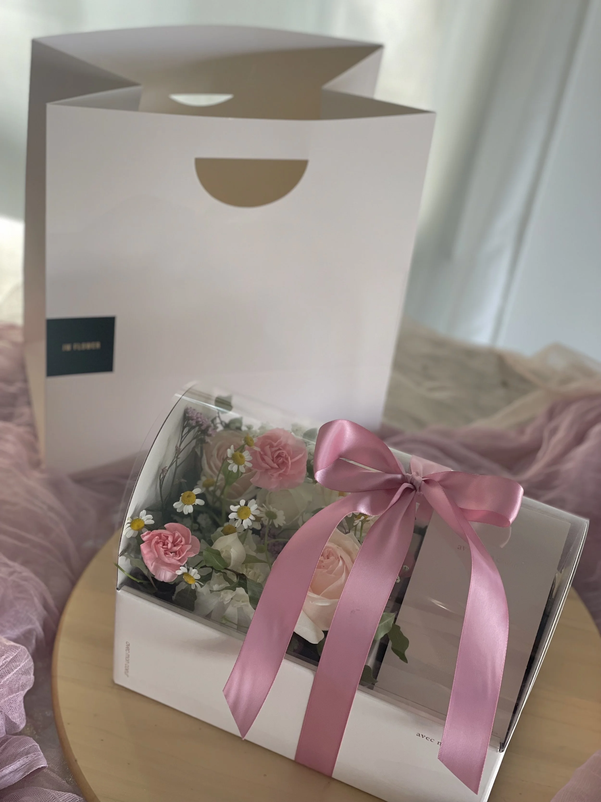 A clear plastic box with pink and white artificial flowers and greenery, decorated with a pink satin ribbon tied in a bow, placed on a wooden surface.
