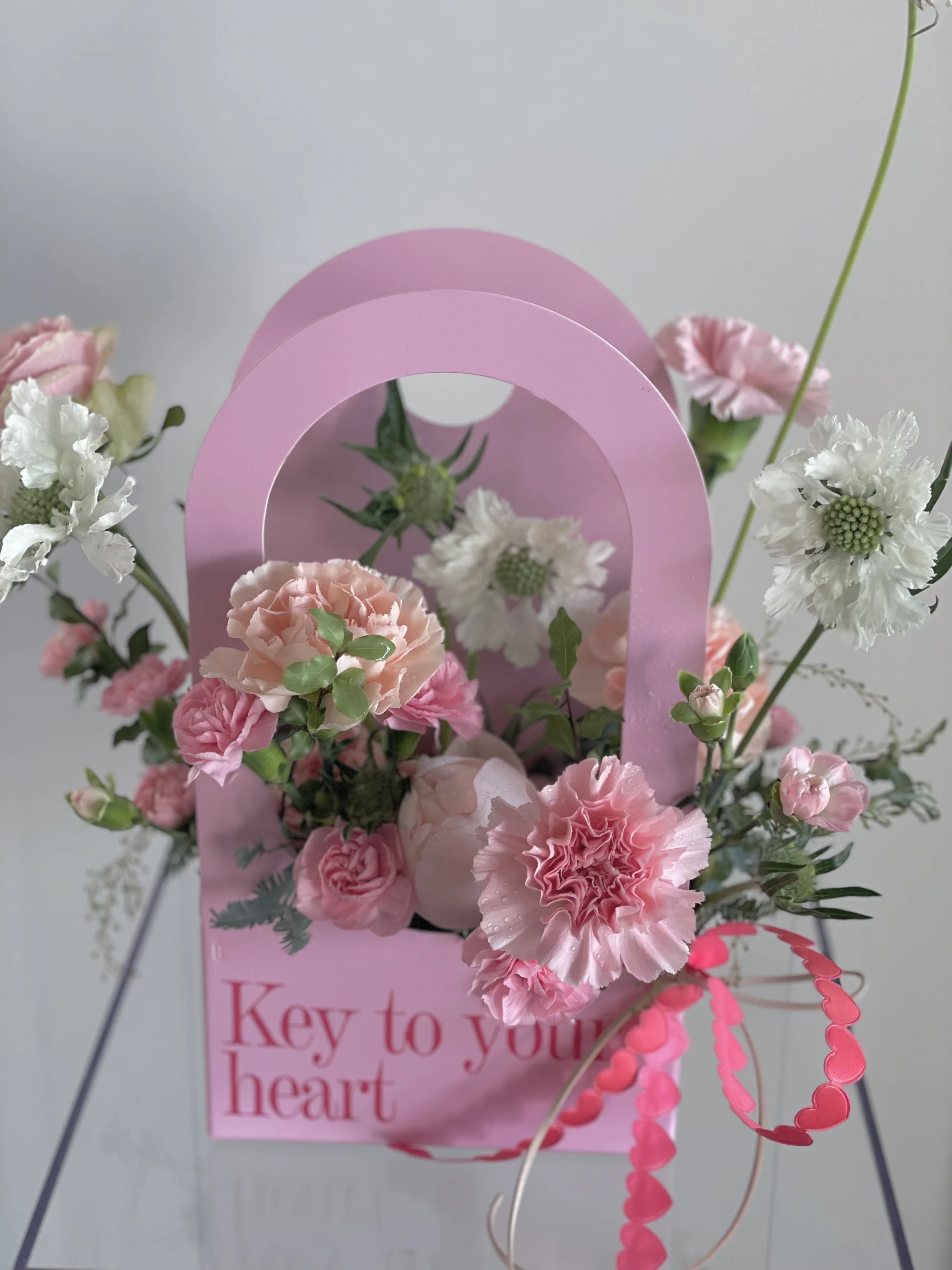 A pink gift bag filled with various pink and white carnations and greenery, with a pink heart-shaped garland on the side and the words "Key to your heart" printed on the bag.