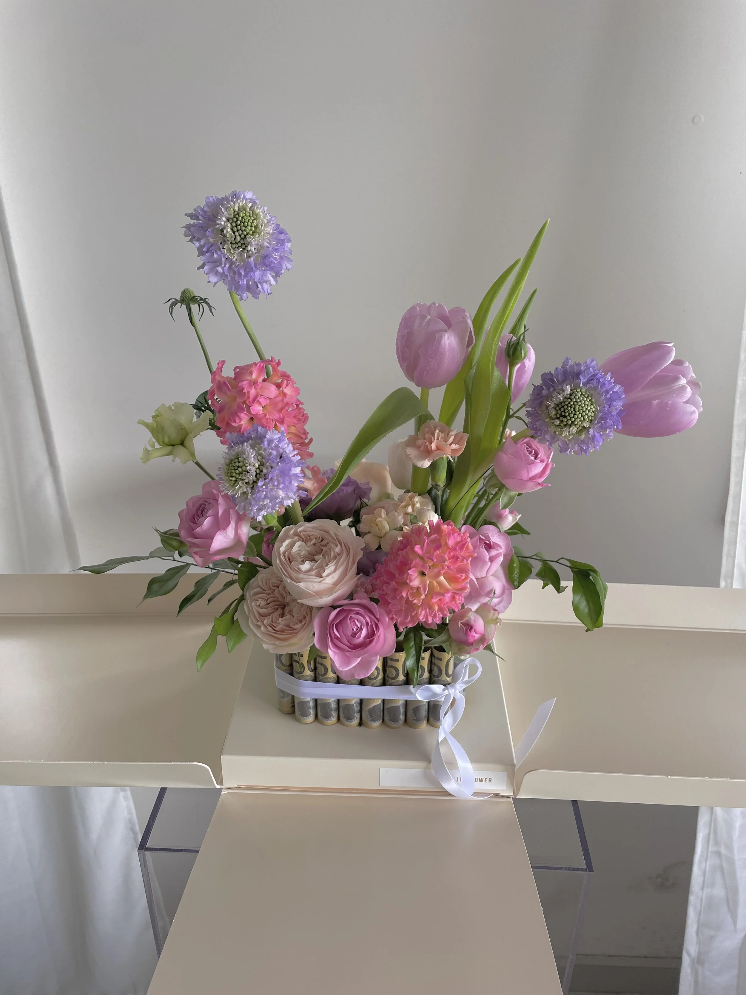 Pink, purple, and peach floral bouquet with tulips, roses, and other flowers in a white vase, placed on a beige table.