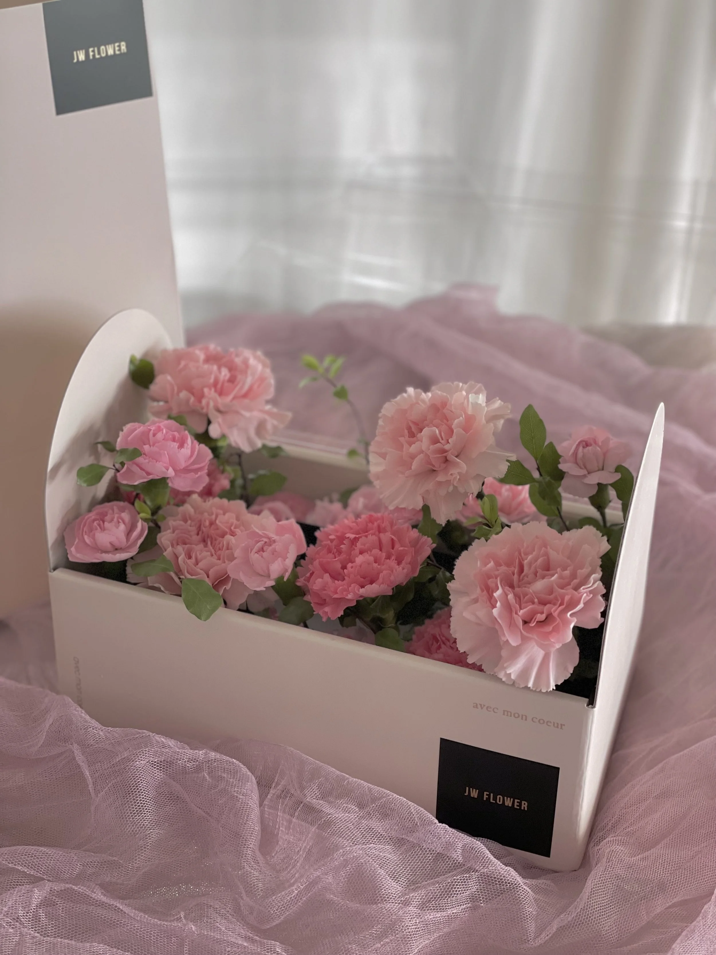 A box of pink carnations and other pink flowers inside a white box with black labels, placed on pink tulle fabric and light-colored background.
