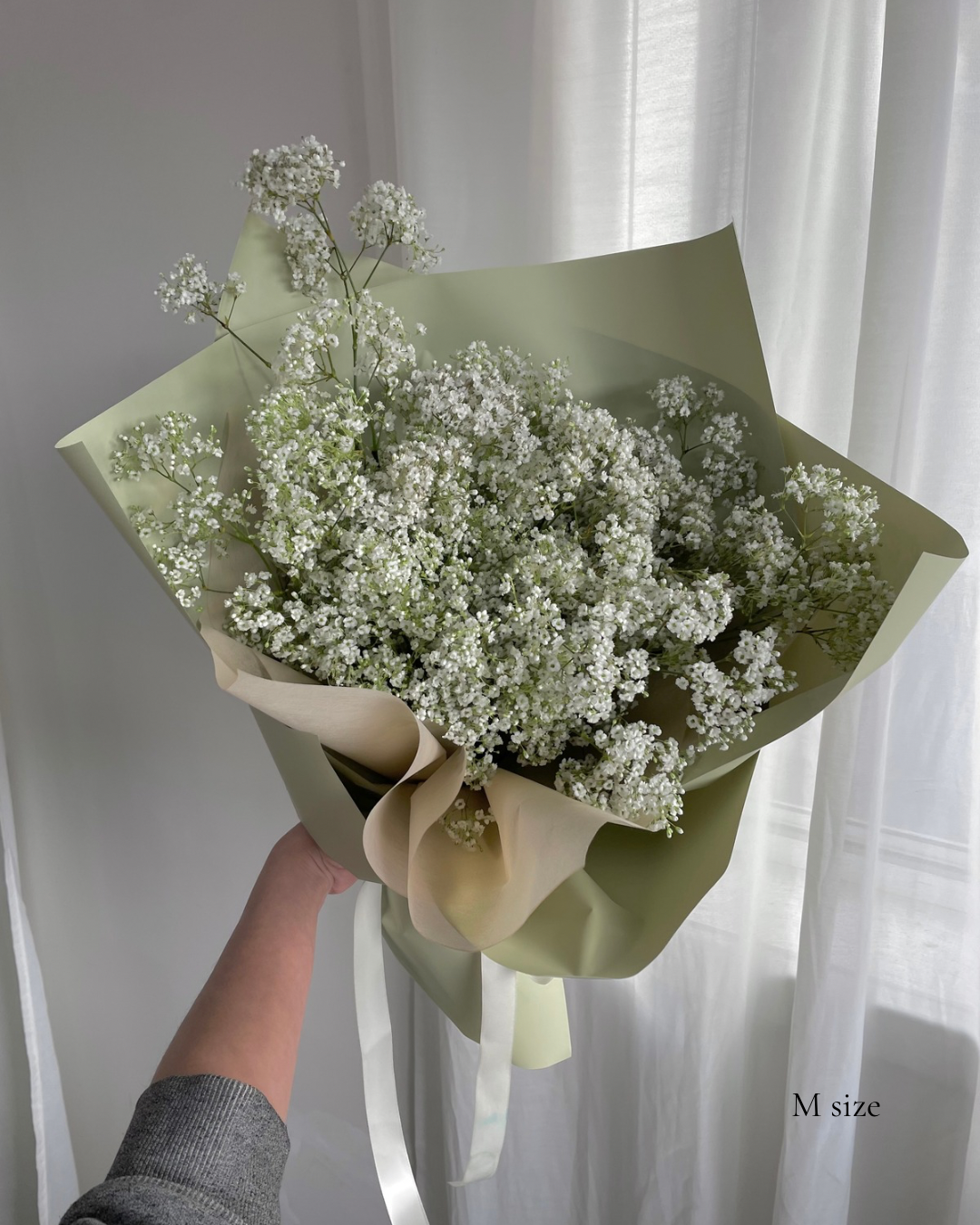 Small bouquet of white baby's breath flowers wrapped in cream and light green wrapping paper, held by a person's hand in front of a white curtain