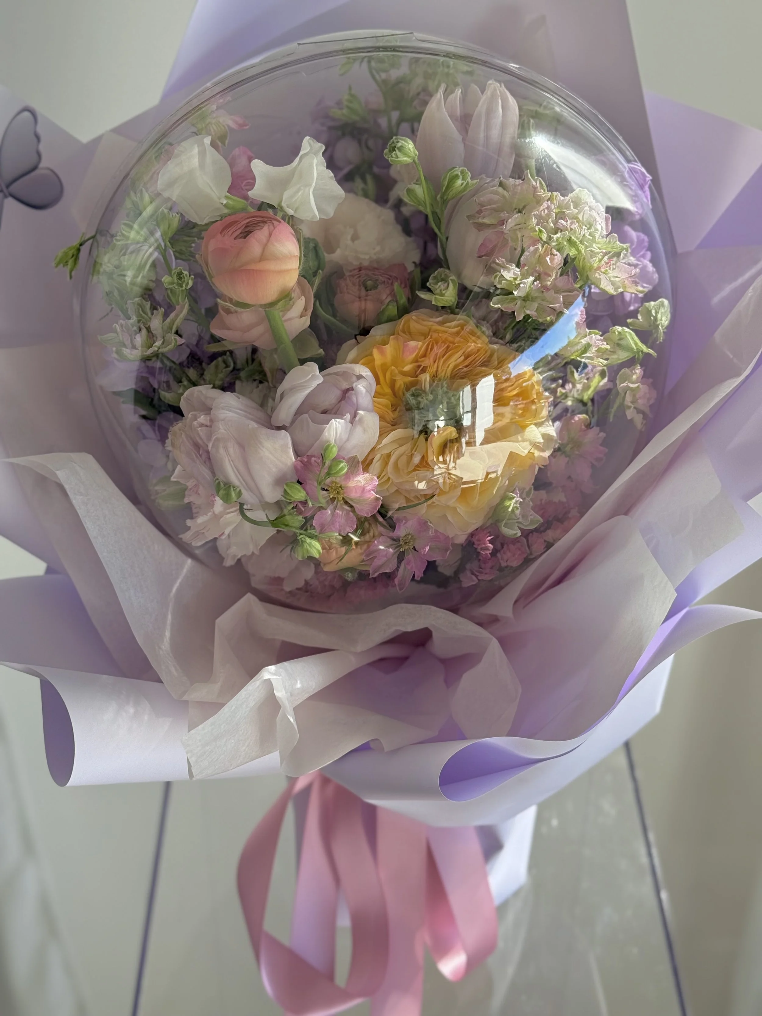 A bouquet of pastel-colored flowers, including roses, tulips, and other blossoms, wrapped in light purple and white paper with pink ribbons.